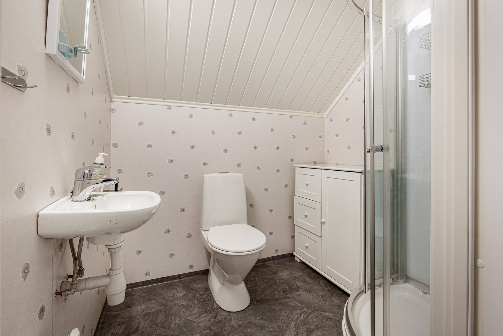 Bad har belegg på gulv med oppbrett på vegg. Vegger med
våtromstapet. Bad har dusjkabinett, vegghengt liten servant og gulvmontert standard toalett Galleribilde