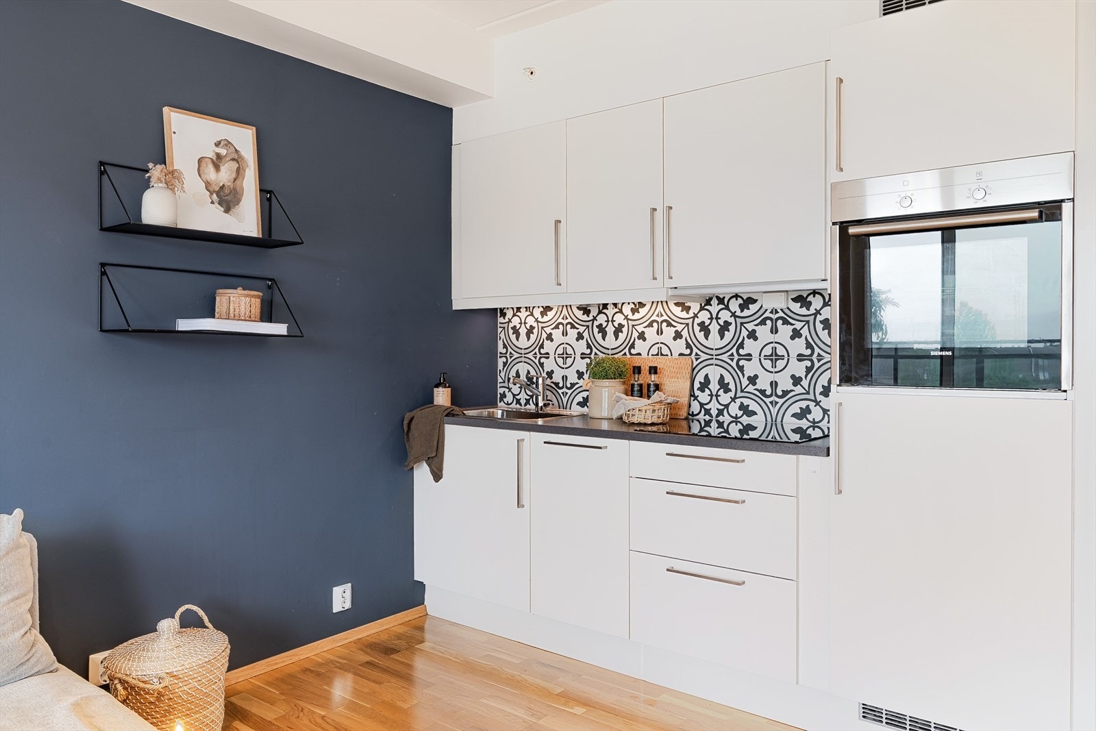 Kjøkkeninnredningen har et enkelt og moderne uttrykk med hvite, slette fronter og mørk benkeplate i laminat. Det er integrert platetopp, stekeovn og ventilator, og løsningen gir god arbeidsflate og skapplass. Galleribilde