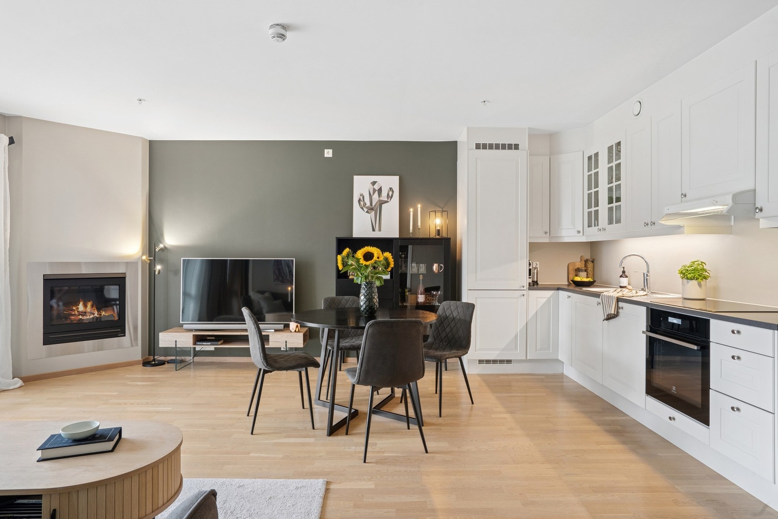 Videre kommer vi inn til en lys og luftig stue i åpen løsning med kjøkken. Her er det plass til både sofakrok og spisebord, og peisen bidrar til en lun atmosfære. Galleribilde