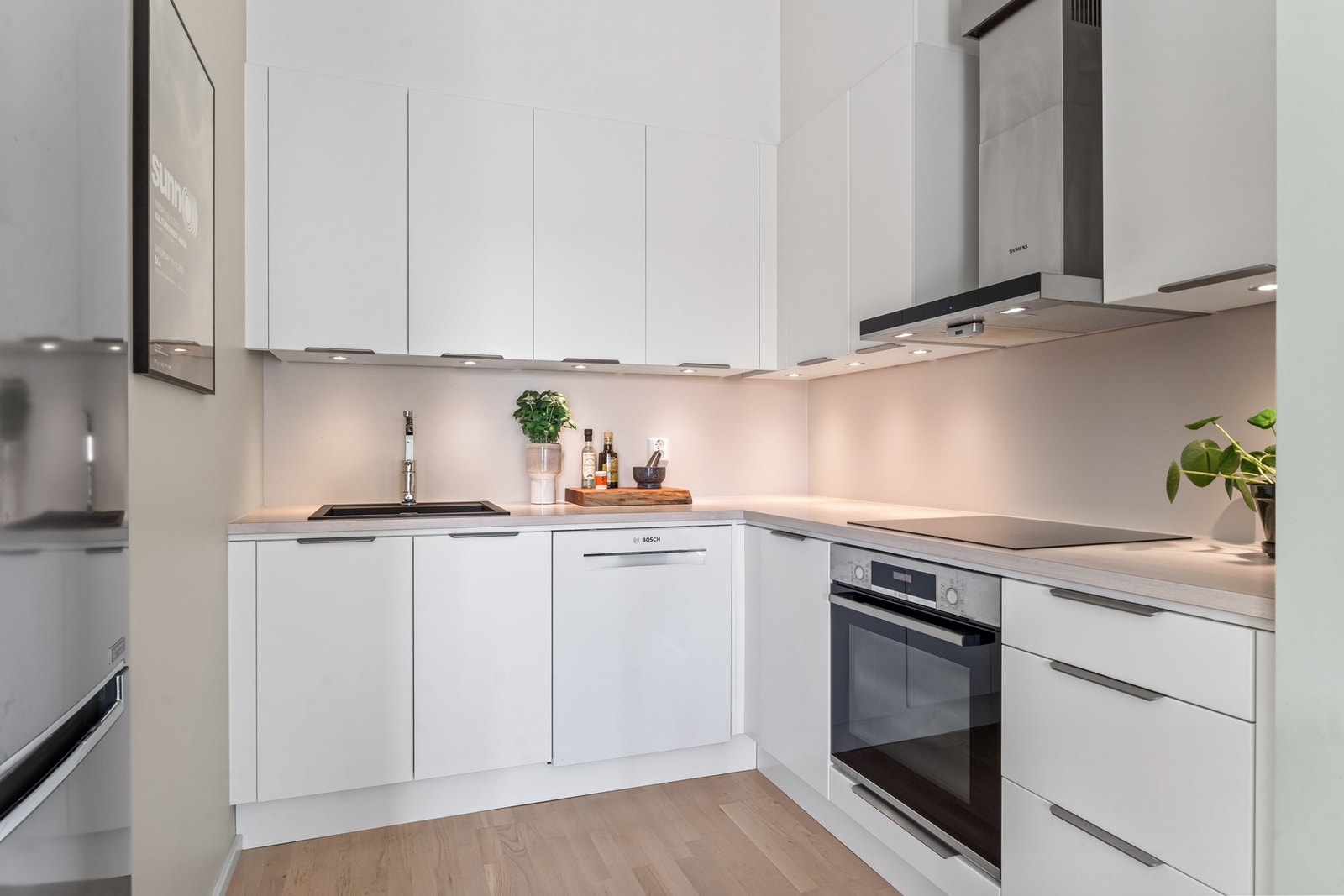 Innredningen er plassert i en L-form, pluss en nisje til kjøleskap langs den tredje veggen. Innredningen har takoppforede skap, hvite, glatte fronter med smale grep, samt benkeplater i grå laminat med nedfelt vaskekum i sort kompositt. Galleribilde