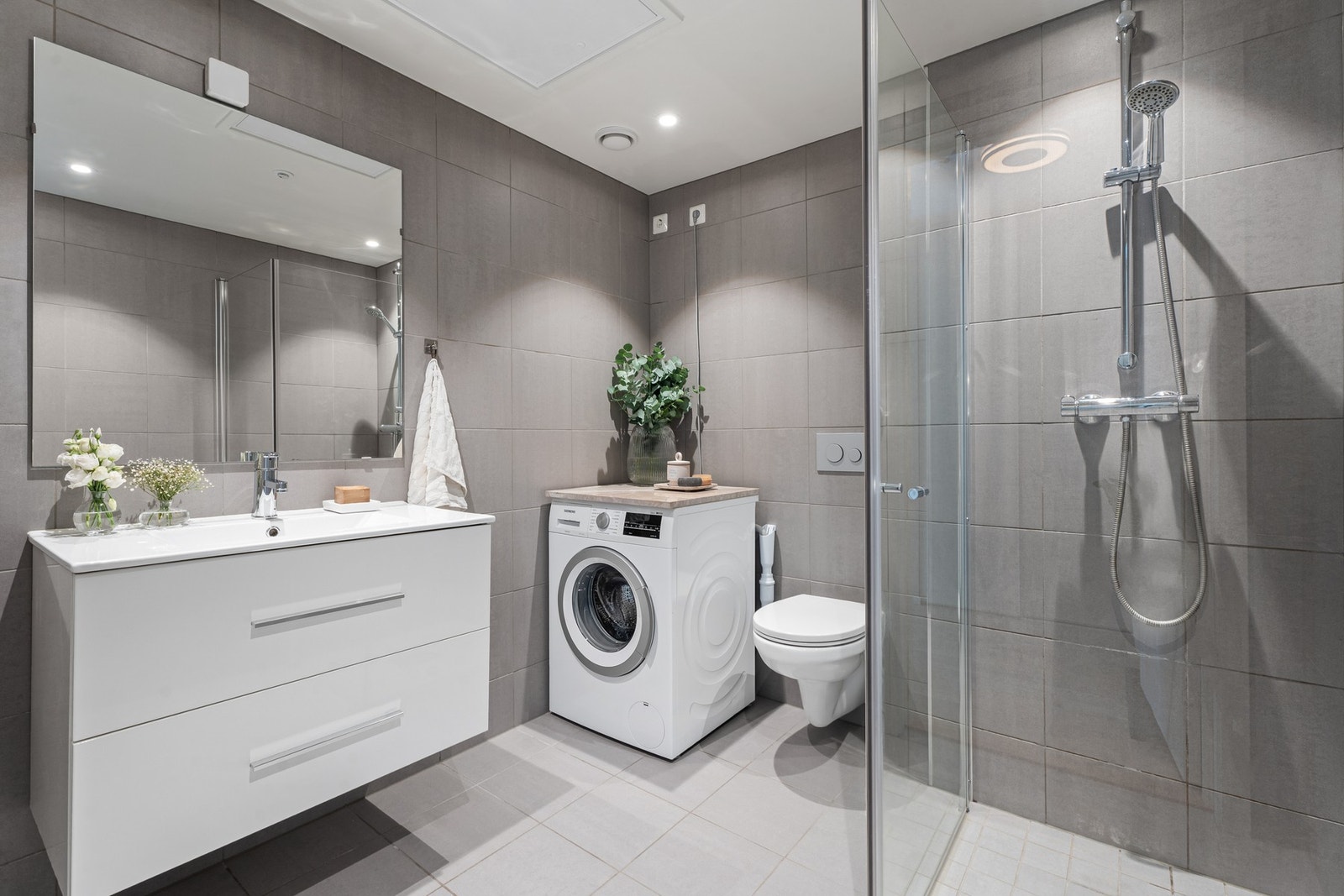 Badet er moderne og pent med veggmontert toalett, dusjhjørne med innfellbare glassdører og opplegg for vaskemaskin. Rommet er utstyrt med varmekabler i gulvet og downlights i himlingen. Galleribilde