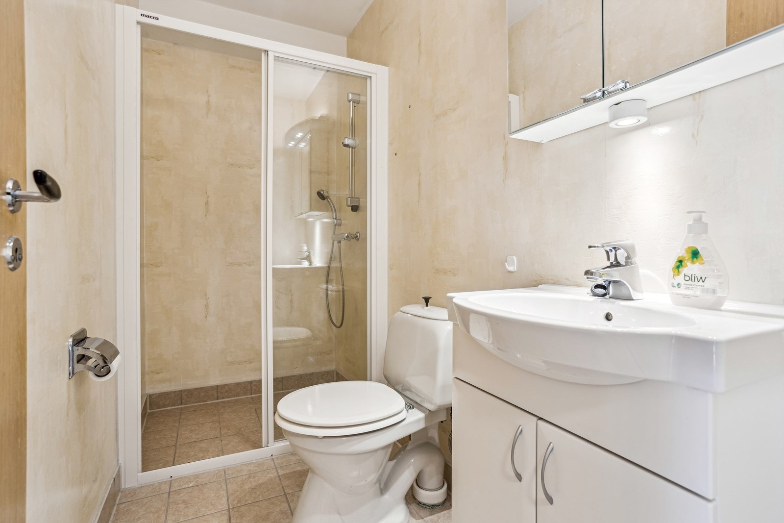 Flislagt gulv med sokkelflis og vinylbelegg på vegger. Badet har servant med skapinnredning, overskap med speilfront, dusjvegg med skyvedør og et toalett. Galleribilde