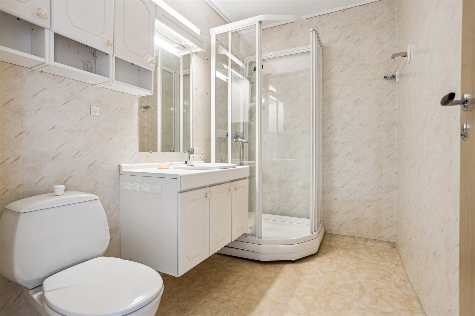 Bad med gulvbelegg og vegger med vinyltapet. Badet er innredet med hvit vask med skapinnredning, overskap og speil, dusjkabinett og et toalett. Galleribilde