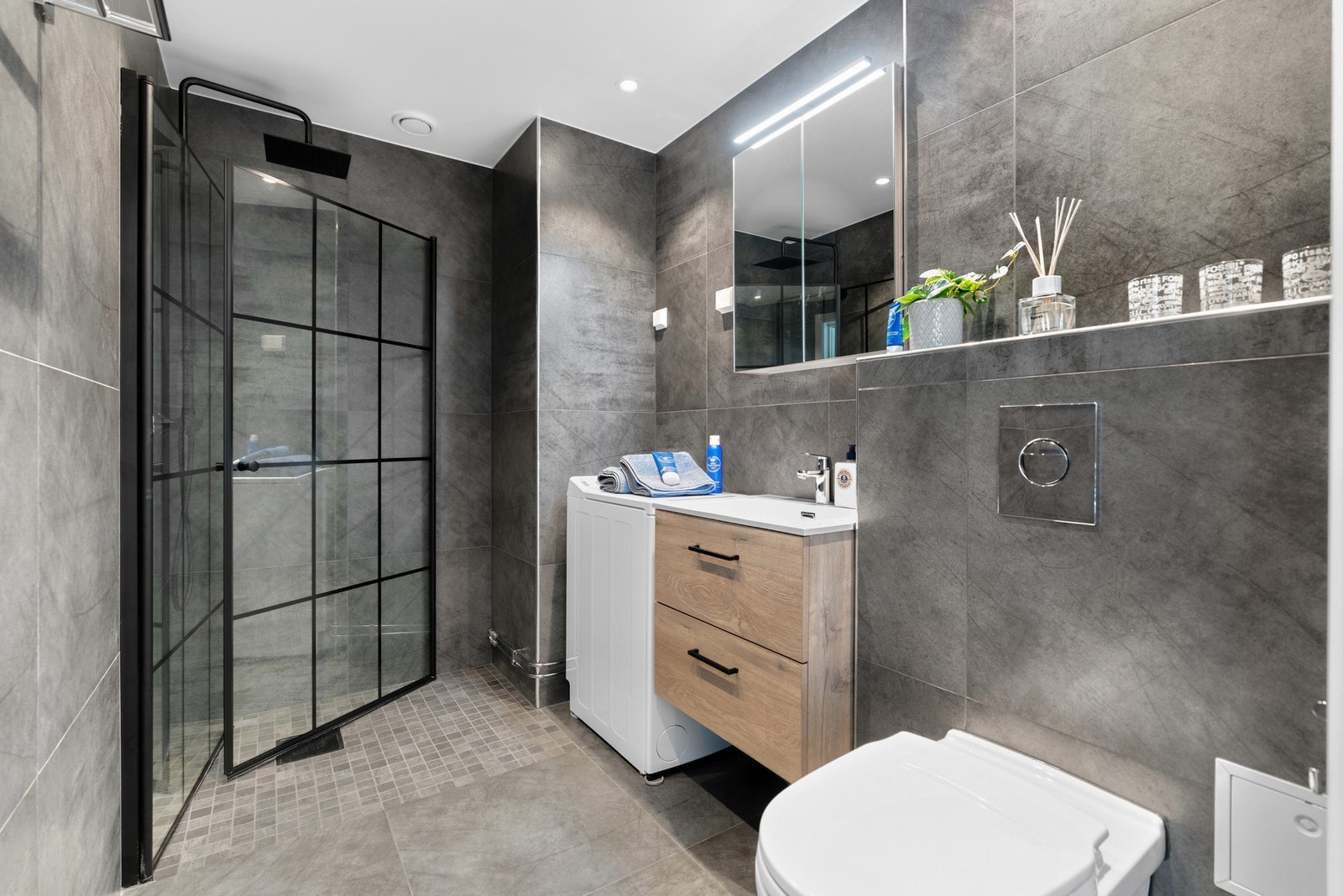 Romslig og delikat baderom fra 2020 med dusjhjørne, innfellbar dusjvegg med sort blandebatteri og sort dusjarmatur, vegghengt toalett og store nydelige fliser på overflater. Galleribilde