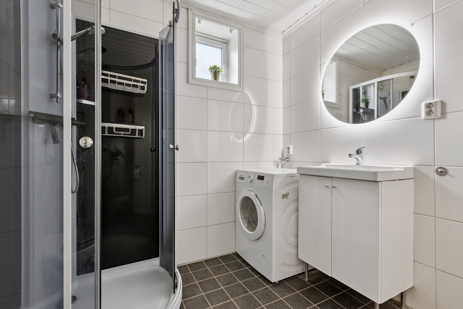 Baderom med flislagt gulv med varme, dusjkabinett, opplegg for vaskemaskin og servant med skap Galleribilde