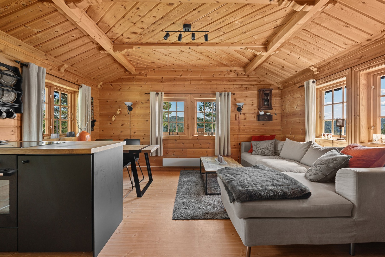 Stue med åpen kjøkkenløsning, store vinduer og trepanel på vegger og tak. Lys og romslig atmosfære. Galleribilde