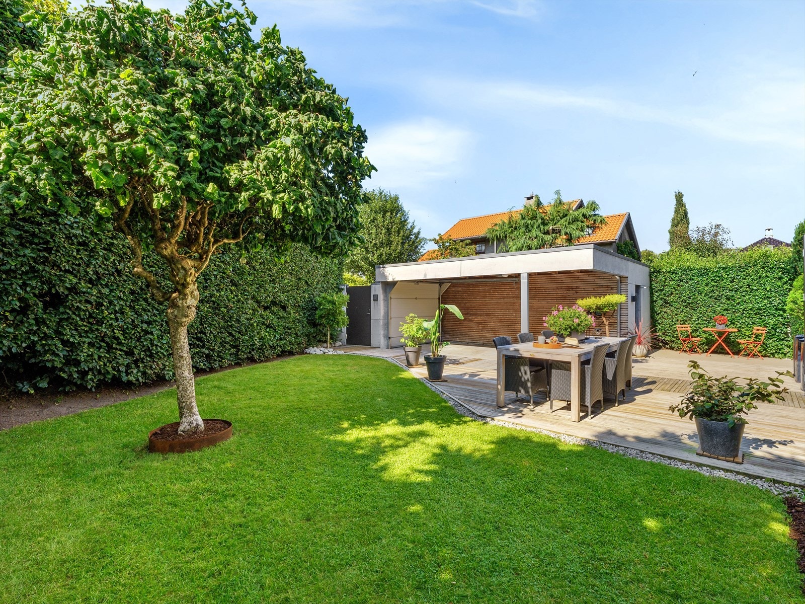 Lune kontraster mellom grønn plen, frodig hekk og trær. Store terrasseplattinger avgrenset av steinbed. Stramkontrast videre til mursteinsvillaen med påbygg. Galleribilde