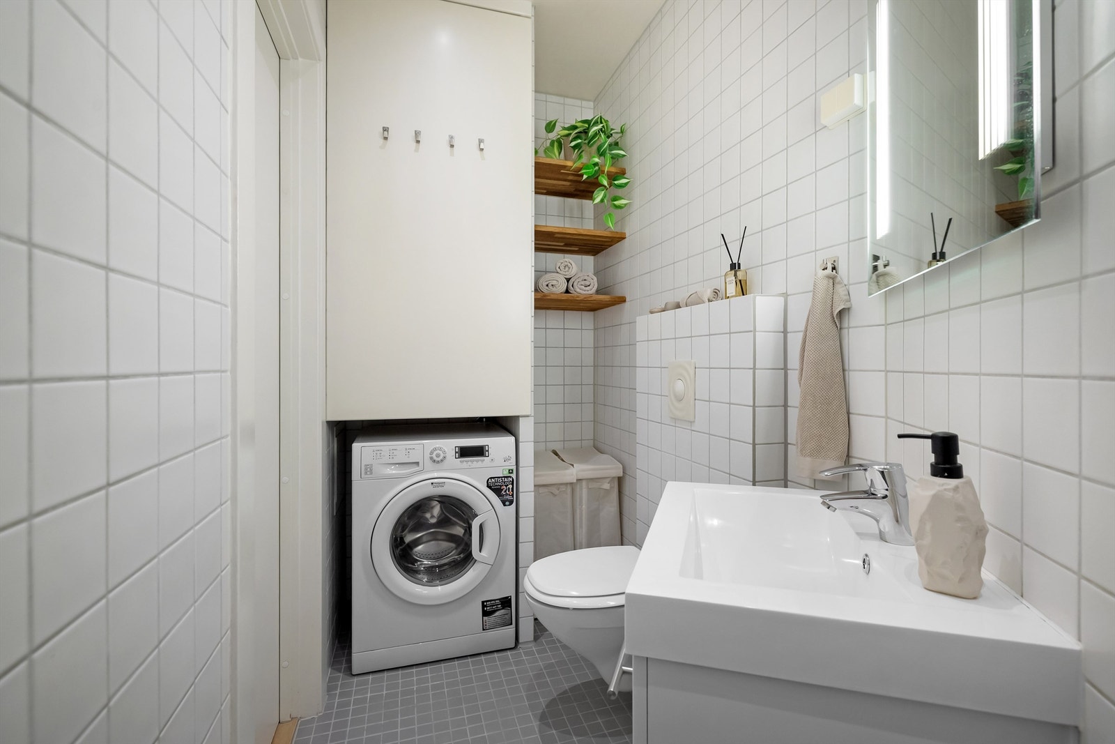 Badet er utstyrt med vegghengt toalett, dusj med dusjforheng, samt opplegg for vaskemaskin og en praktisk nisje med hyller og plass til blant annet skittentøyskurv Galleribilde