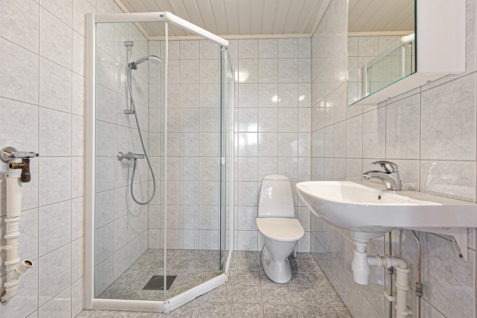 Flislagt baderom med dusjnisje med tett sokkel, servant, toalett og opplegg for vaskemaskin. Galleribilde