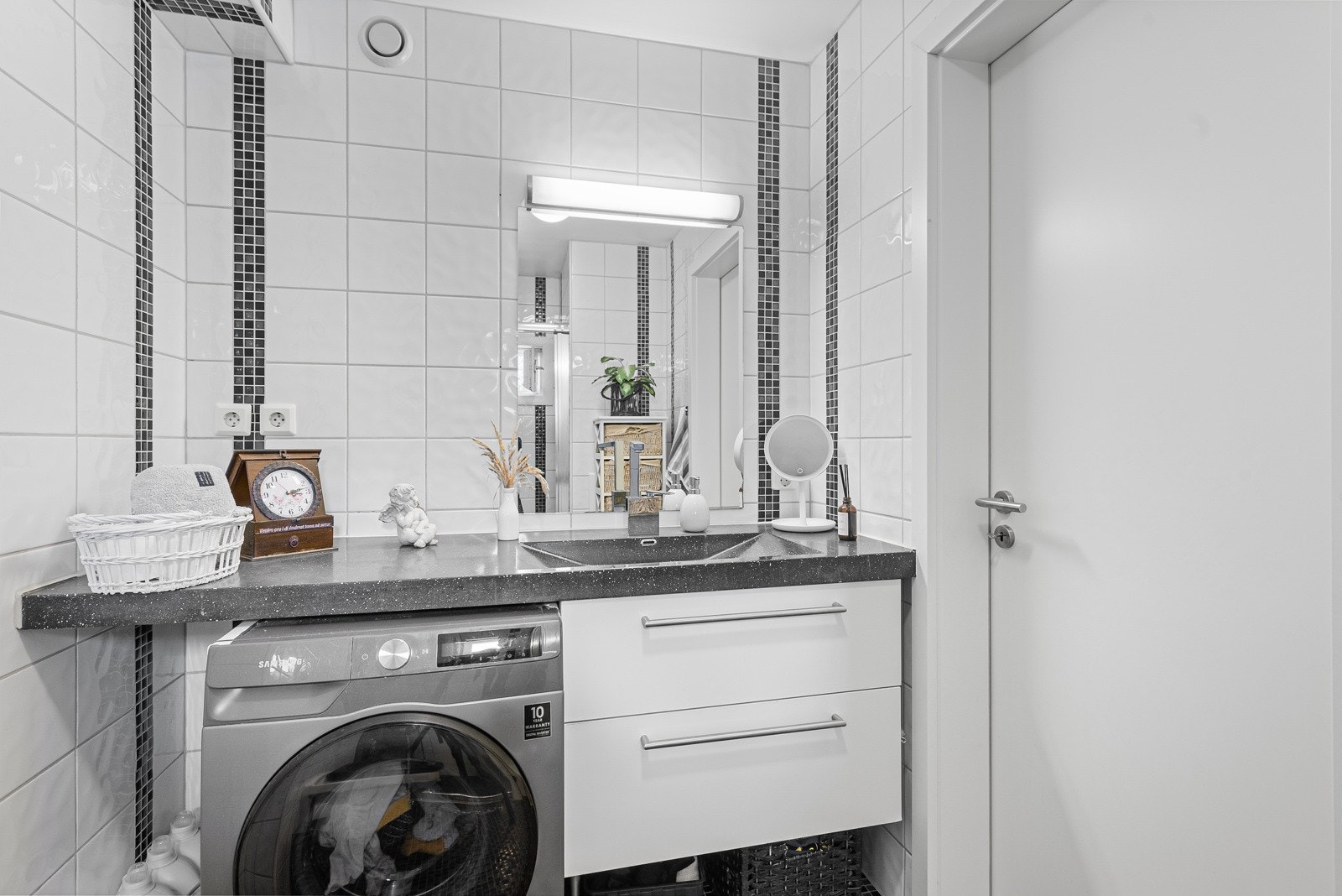 Leiligeten har et tidløst pent bad. Langs den ene veggen har badet en benkeplate med hvite skuffer og nedfelt servant - plassert under et speil med belysning. Under benken er det opplegg for vaskemaskin. Galleribilde