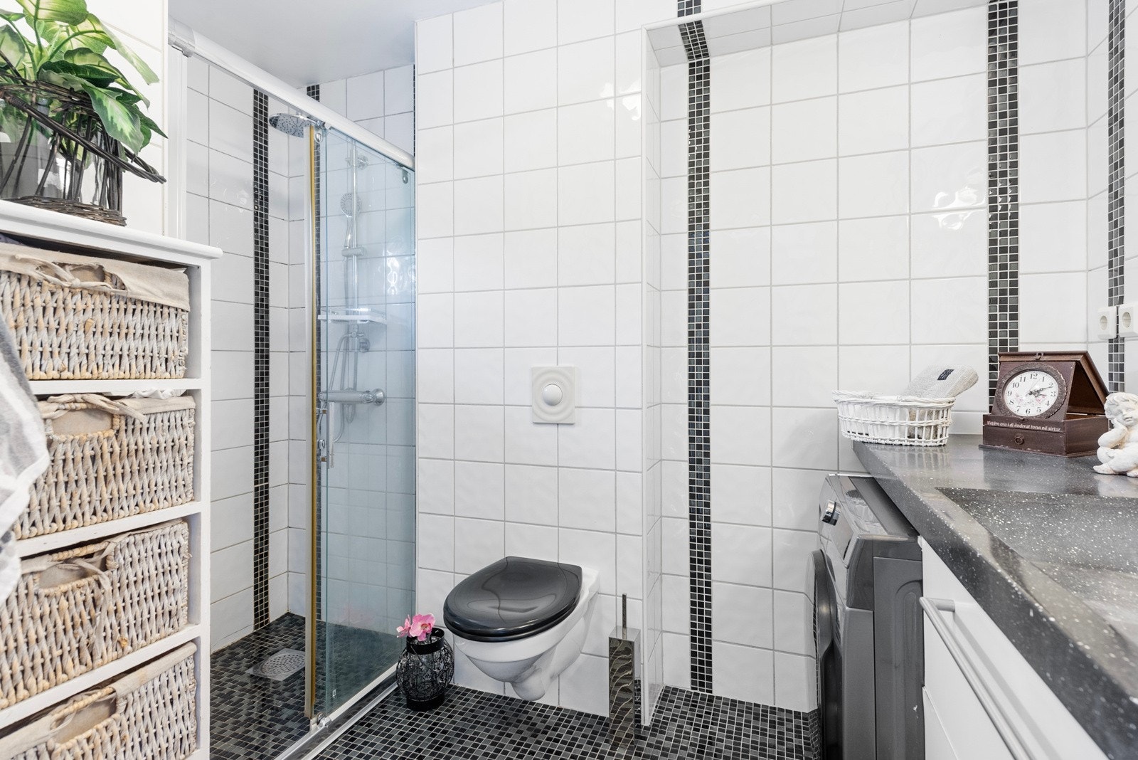 Badet har varmekabler og vegghengt toalett, og et dusjhjørne er utstyrt med skyvedører og regnfallsdusj. Mørke mosaikkfliser på gulvet kombineres med hvite fliser og innslag av mosaikk på veggene. Galleribilde