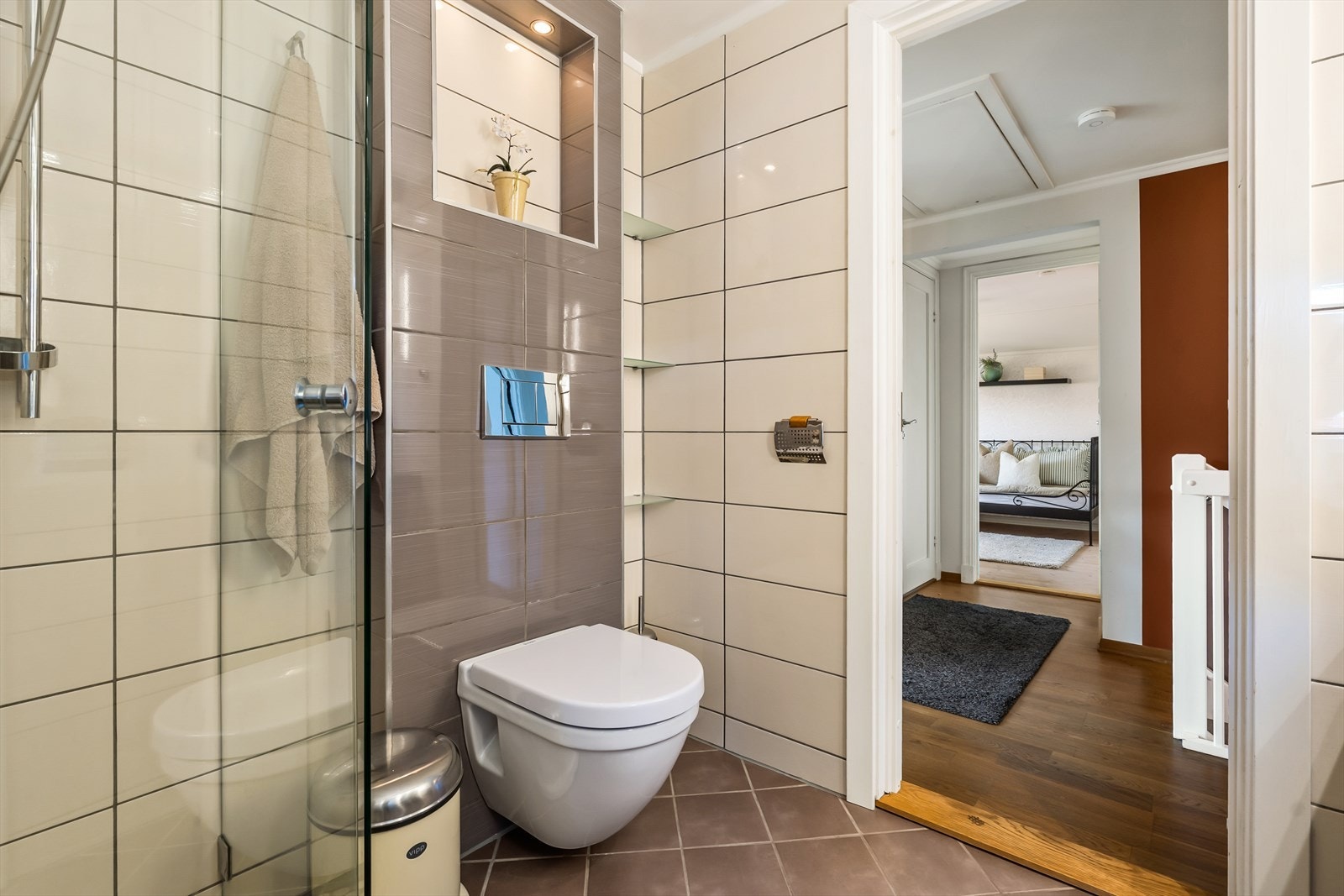 Badet inneholder vegghengt toalett, dusjhjørne med innfellbar glassdør og bred baderomsinnredning med benkeplate med overflate av flis, bolleservant, ettgreps armatur, speil og hyller. Galleribilde