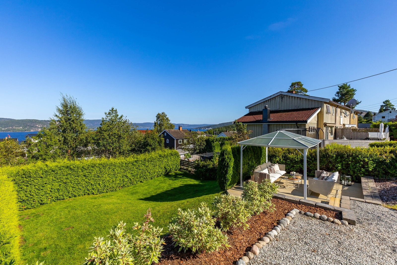 Uteplass med overbygd sittegruppe, plen og beplantning. Utsikt mot sjøen og omkringliggende bebyggelse. Galleribilde