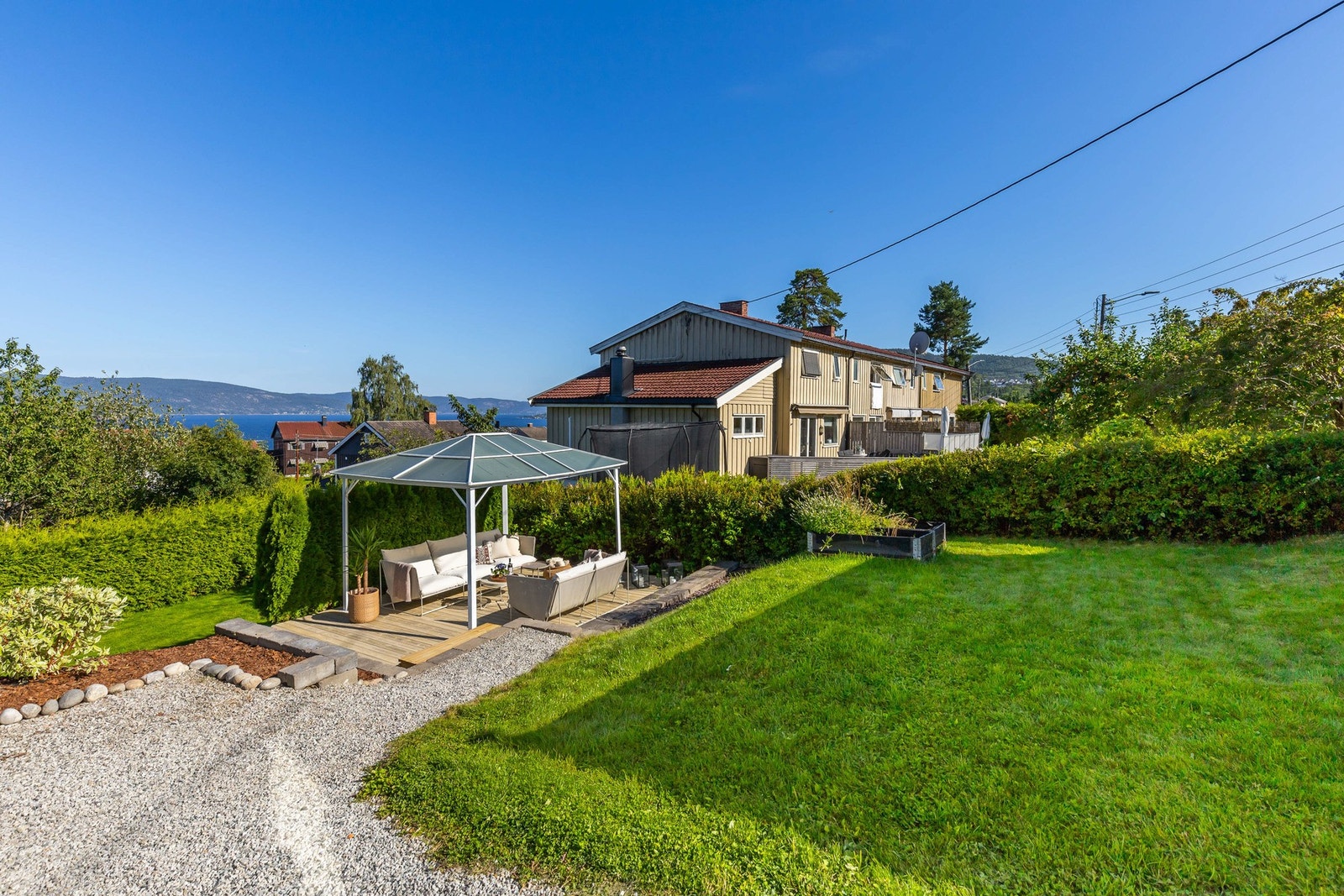 Stor hage med grønt gress, gruslagt sti og sitteområde under pergola. Galleribilde