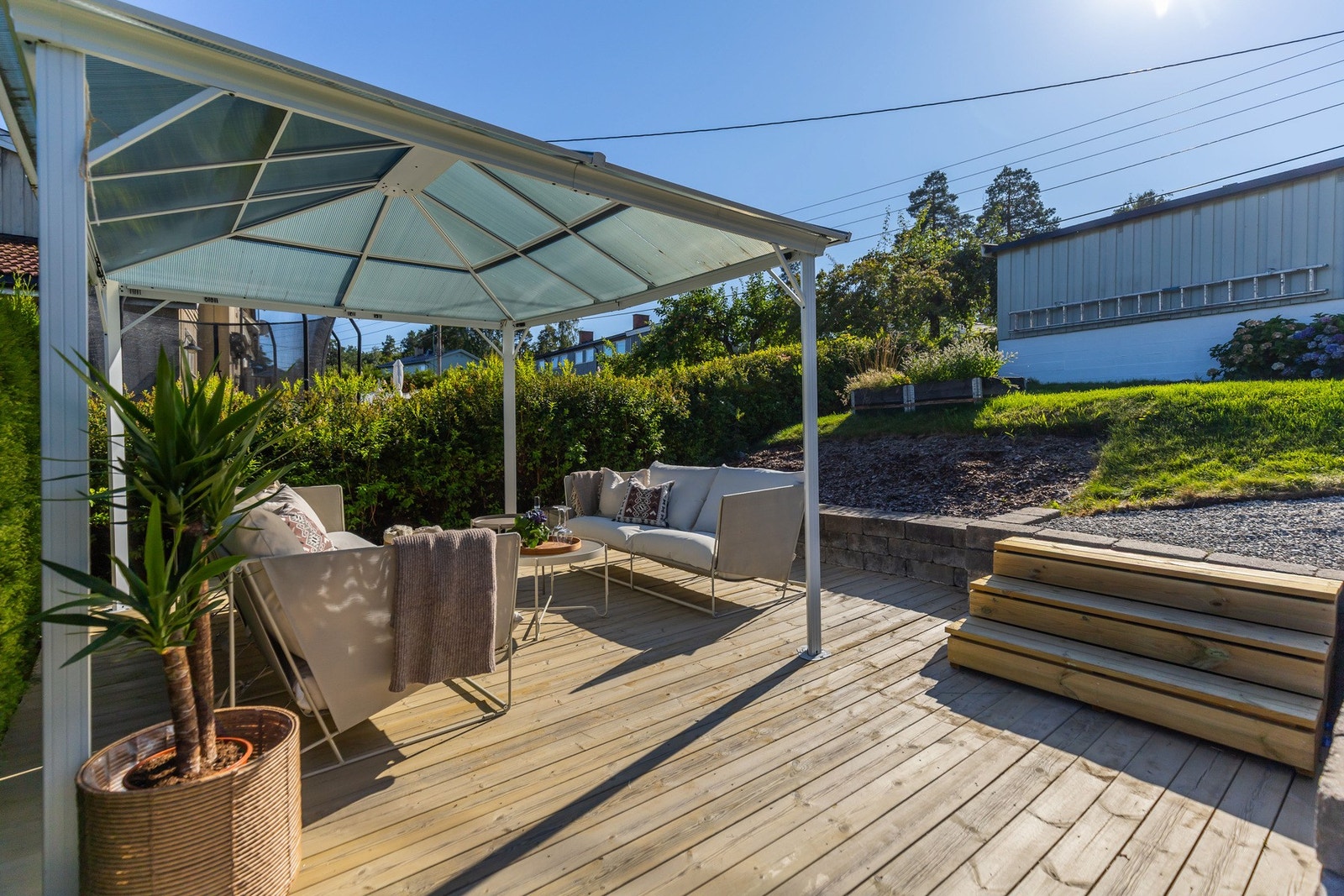 Overbygd uteplass med sittegruppe på treterrasse, omgitt av grøntområder og gruslagt gangvei. Galleribilde