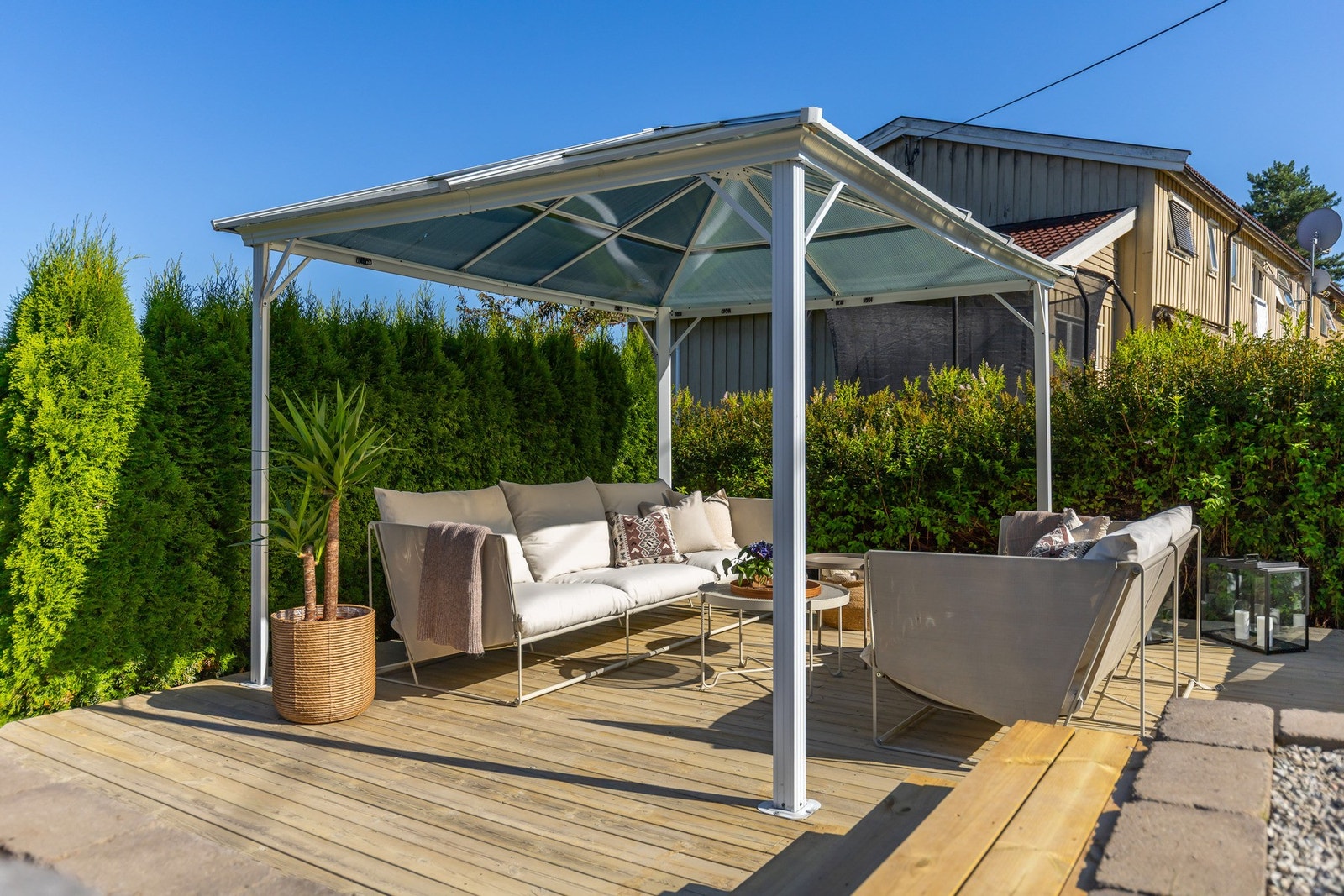 Utendørs sittegruppe under tak på treterrasse, omgitt av grønne busker og trær. Galleribilde