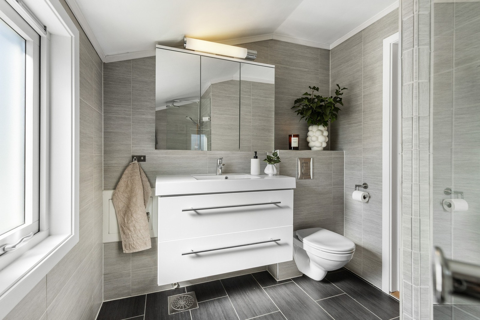 Moderne bad med grå fliser, vegghengt toalett og speilskap over servant. Galleribilde