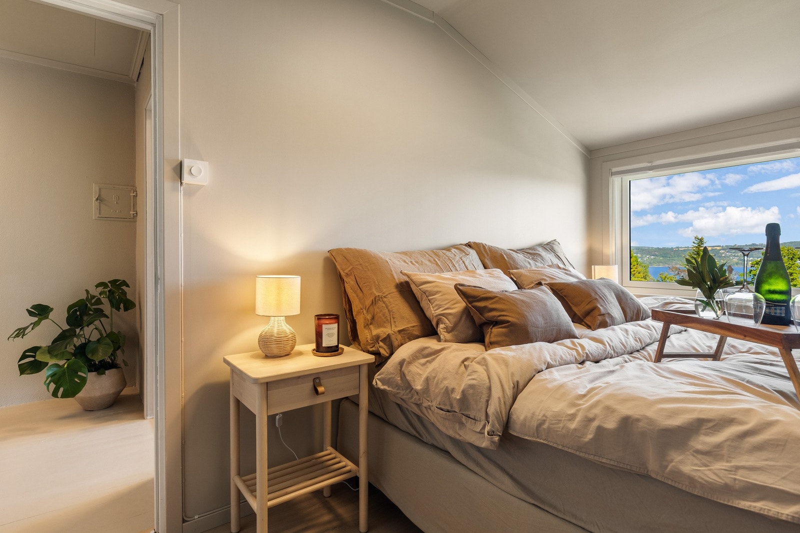 Lyst soverom med stor seng, nattbord og vindu med utsikt over fjorden og grønne åser. Galleribilde