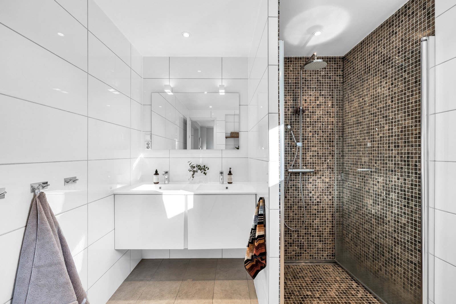 Badet har brune gulvfliser og hvite veggfliser, mens dusjnisjen har mosaikk i lekre farger. Galleribilde