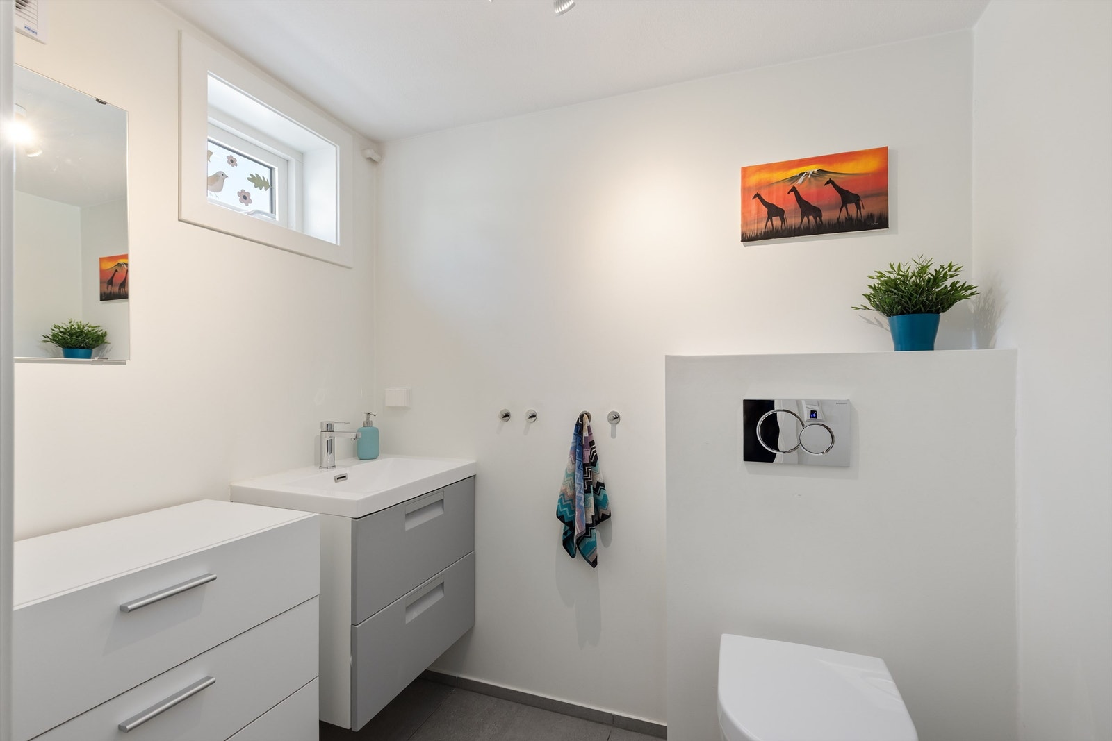 Toalettrom i kjeller med flislagt gulv med gulvvarme, vegghengt toalett og servant med underliggende servantskuffer Galleribilde