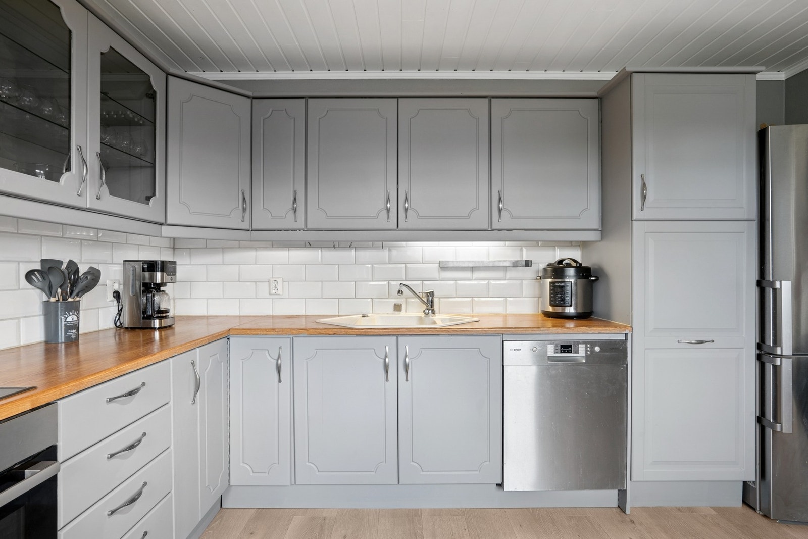 Den tidløse innredningen har profilerte fronter i grått, kombinert med lyse benkeplater og klassisk hvit flisvegg. Galleribilde