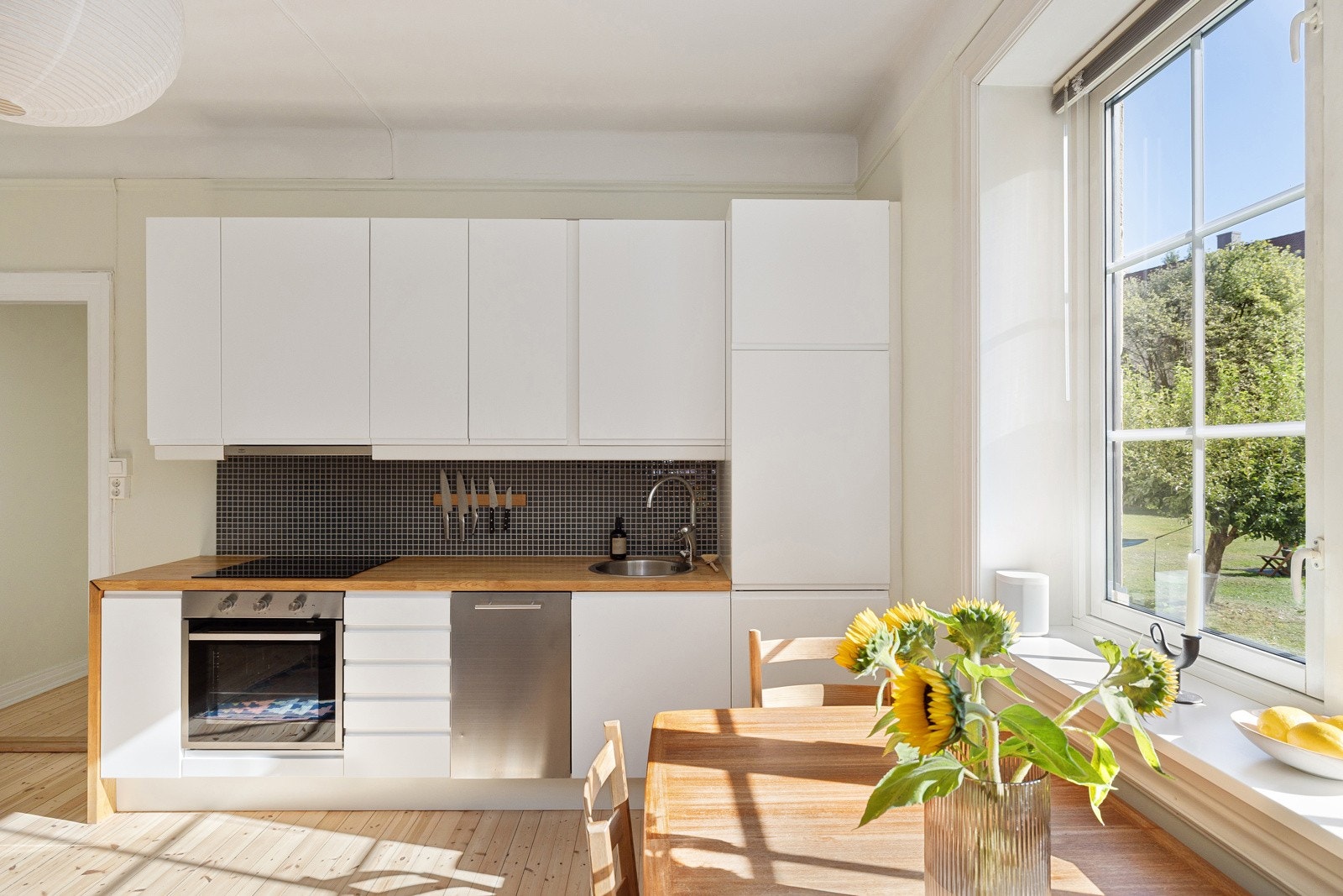 Lys og pen kjøkkeninnredning med god skap- og benkeplass. Innredningen har hvite glatte fronter og benkeplate av tre med nedfelt oppvaskkum. Både fint og praktisk med mosaikkfliser i benkeryggen og belysning under overskapene. Galleribilde
