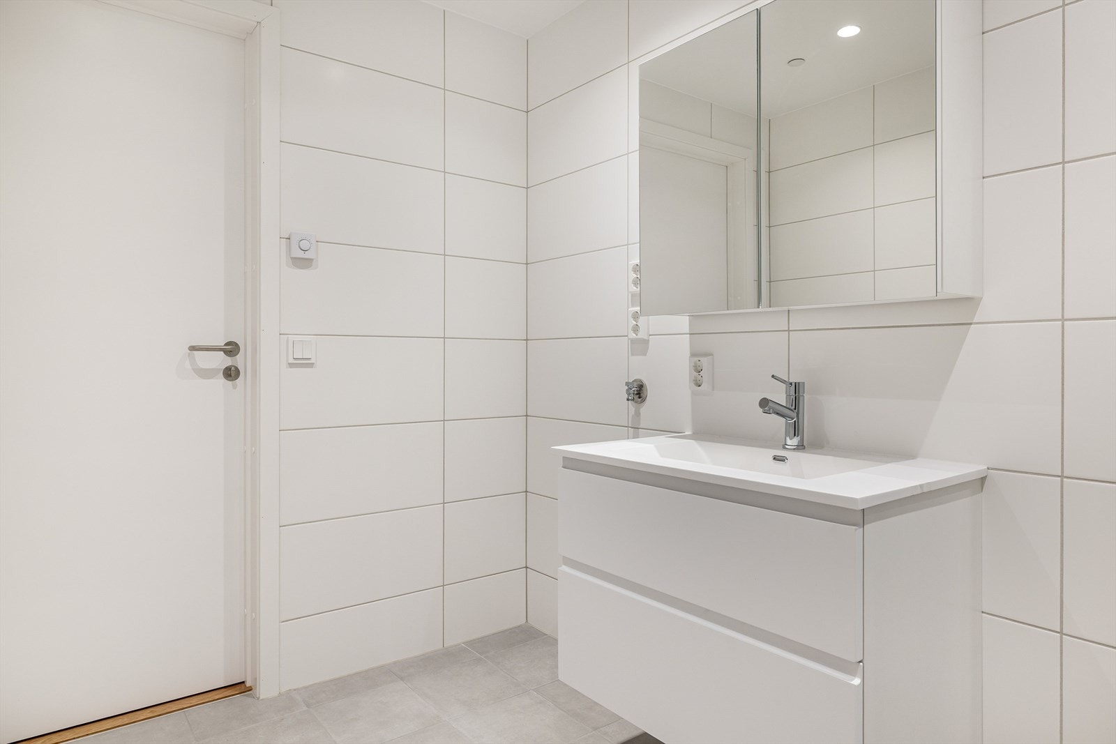 Moderne baderomsinnredning med skuffer og heldekkende servant, samt vegghengt speilskap over. I hjørnet er det plass og opplegg for vaskemaskin. Galleribilde