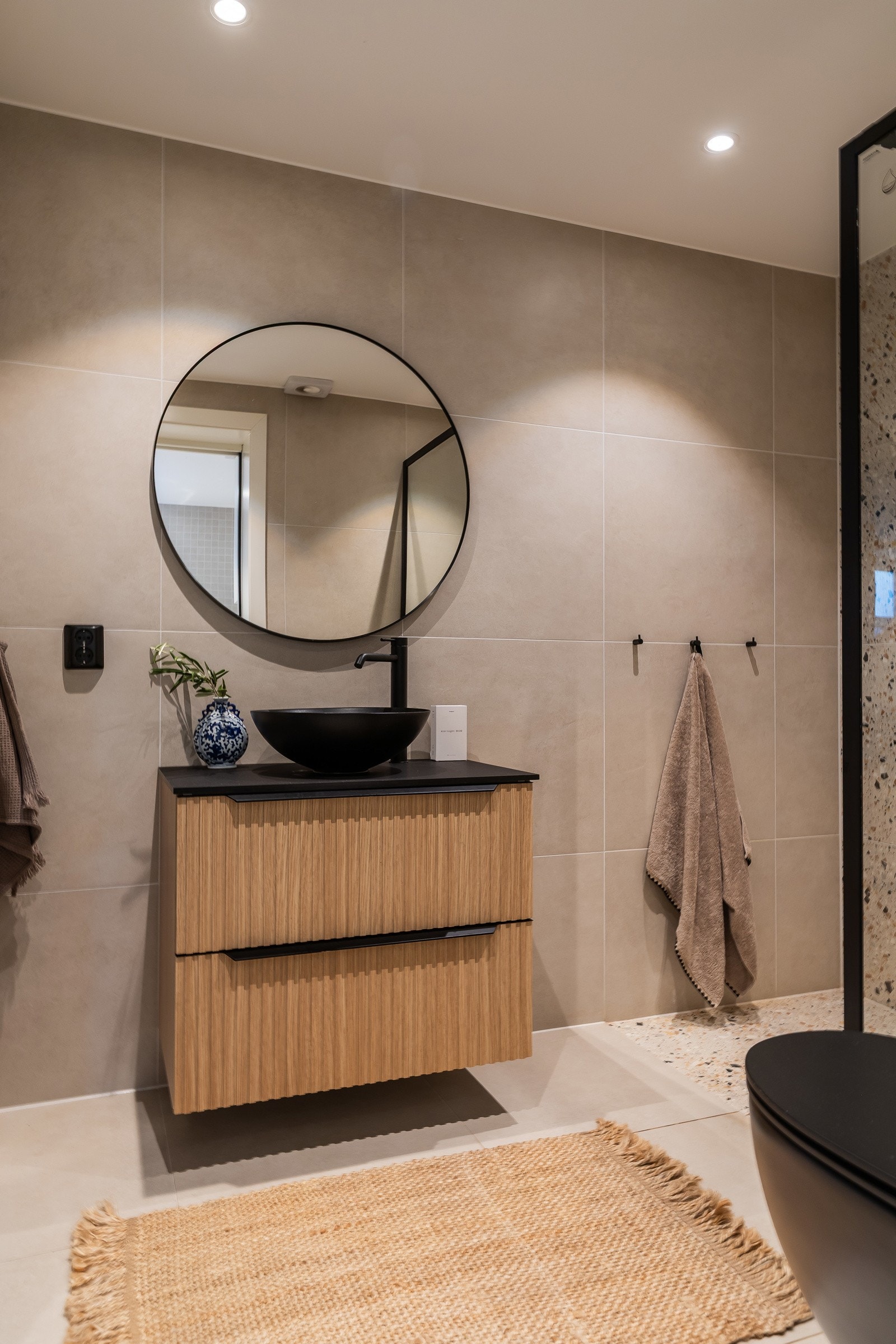 Stilig baderomsinnredning, med skuffer og ovenpåliggende servant med ett-greps armatur. Stort rundt veggspeil. Baderommet er også utstyrt med dusjhjørne med glassvegg og ett-greps armatur med dusjhode i tillegg til regnfallsdusj samt vegghengt toalett. Galleribilde