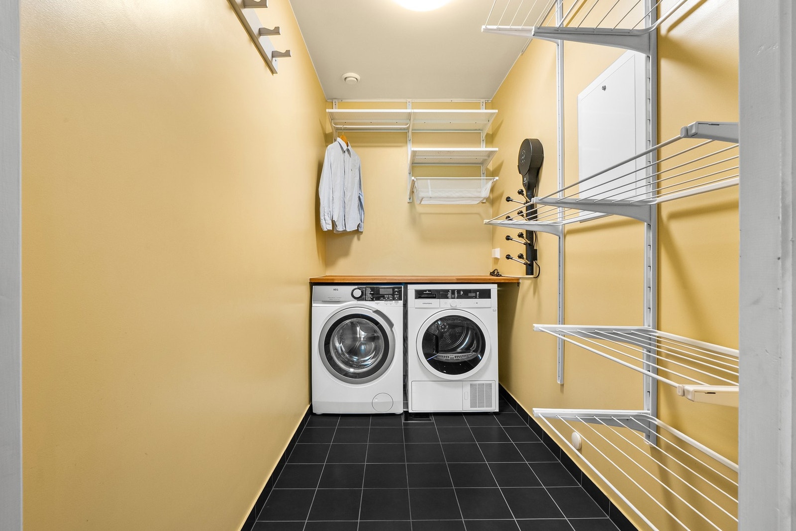 Vaskerom med vaskemaskin og tørketrommel, flislagt gulv og flere hyller for oppbevaring. Galleribilde