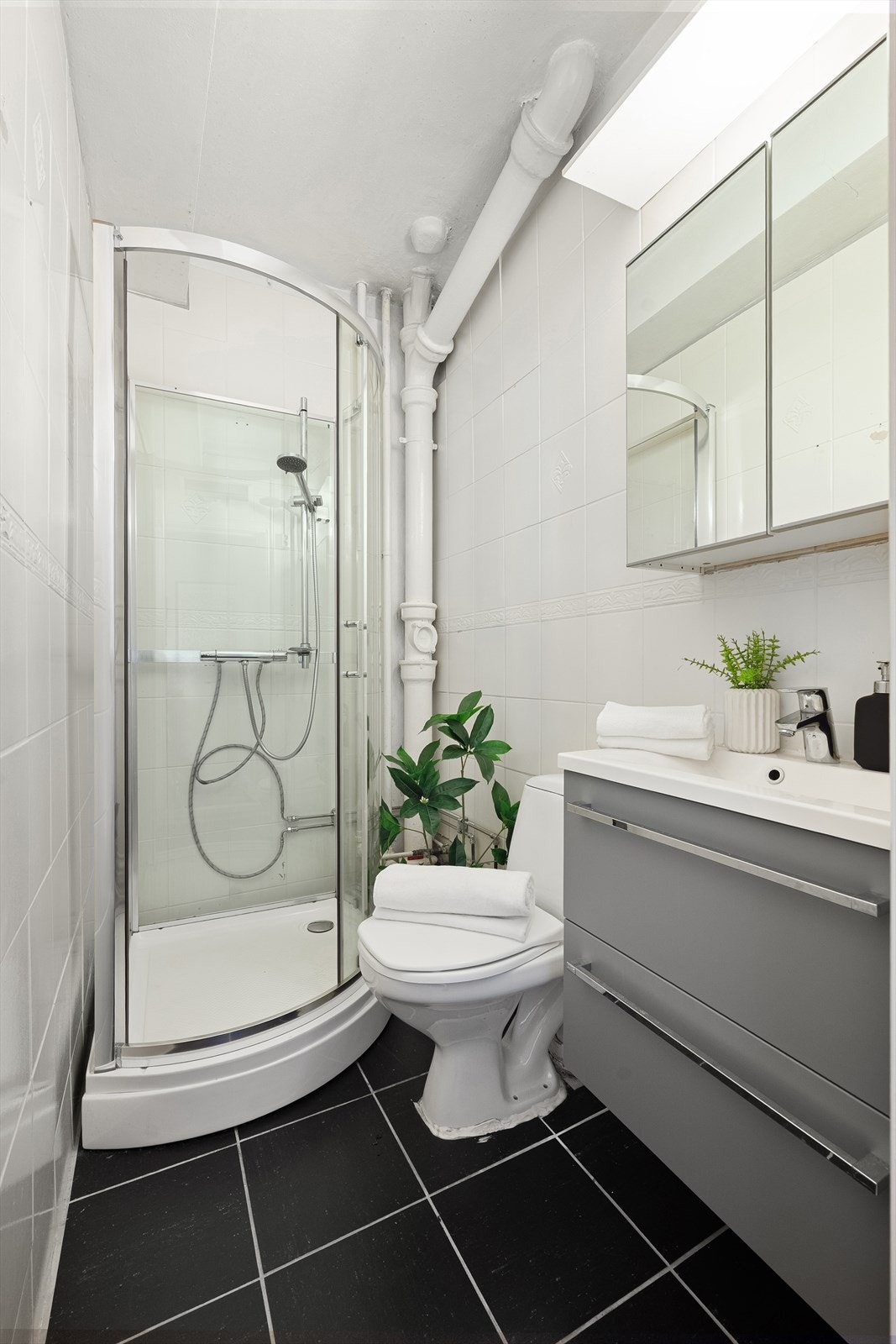 Badet er innredet med mørke fliser på gulvet, lyse fliser på veggene og ellers dusjkabinett, toalett, servant i skuffeinnredning og speilskap for praktisk oppbevaring. Galleribilde