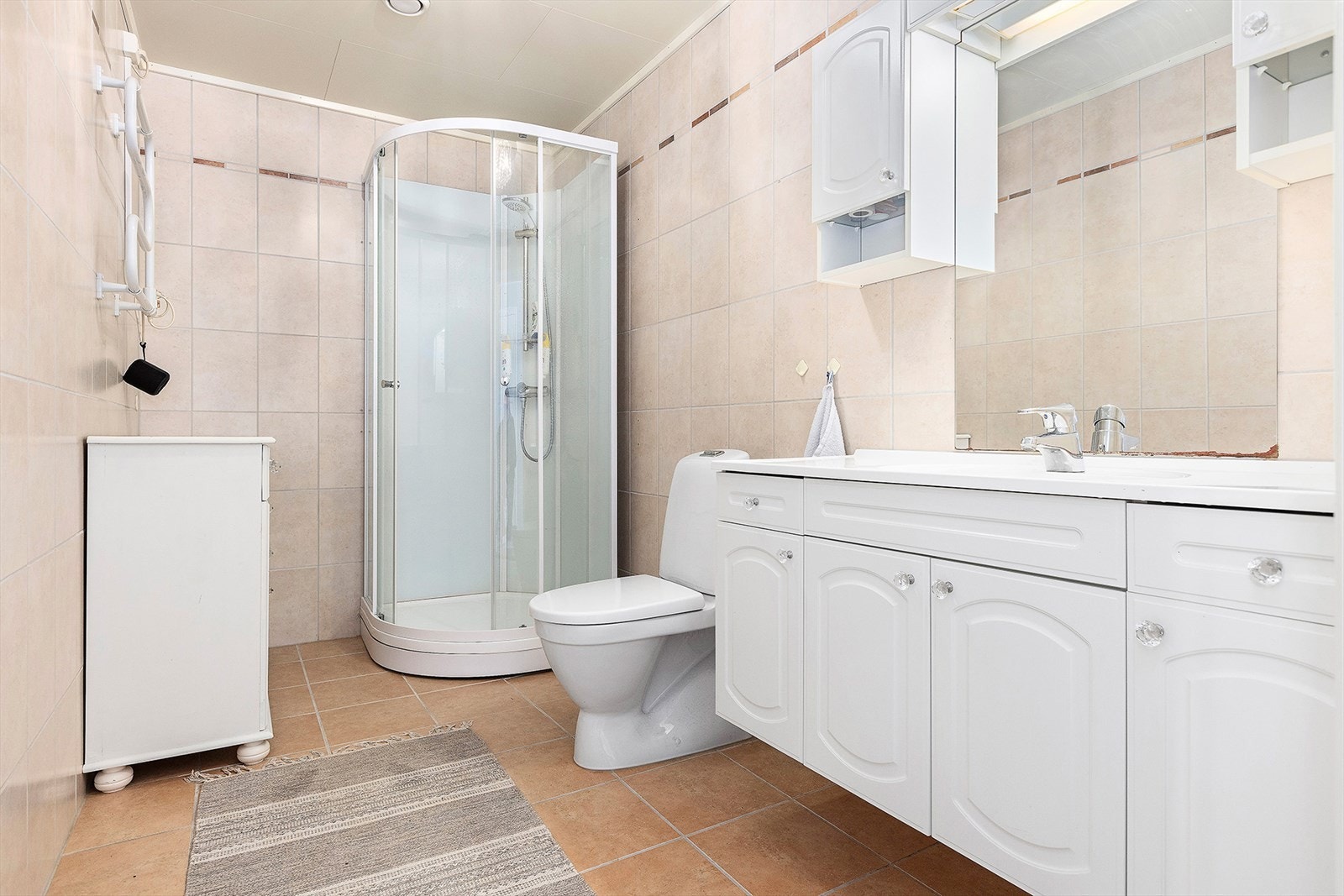 Bad med flis på gulv og vegg. Montert dusjkabinett, toalett og innredning med servant Galleribilde