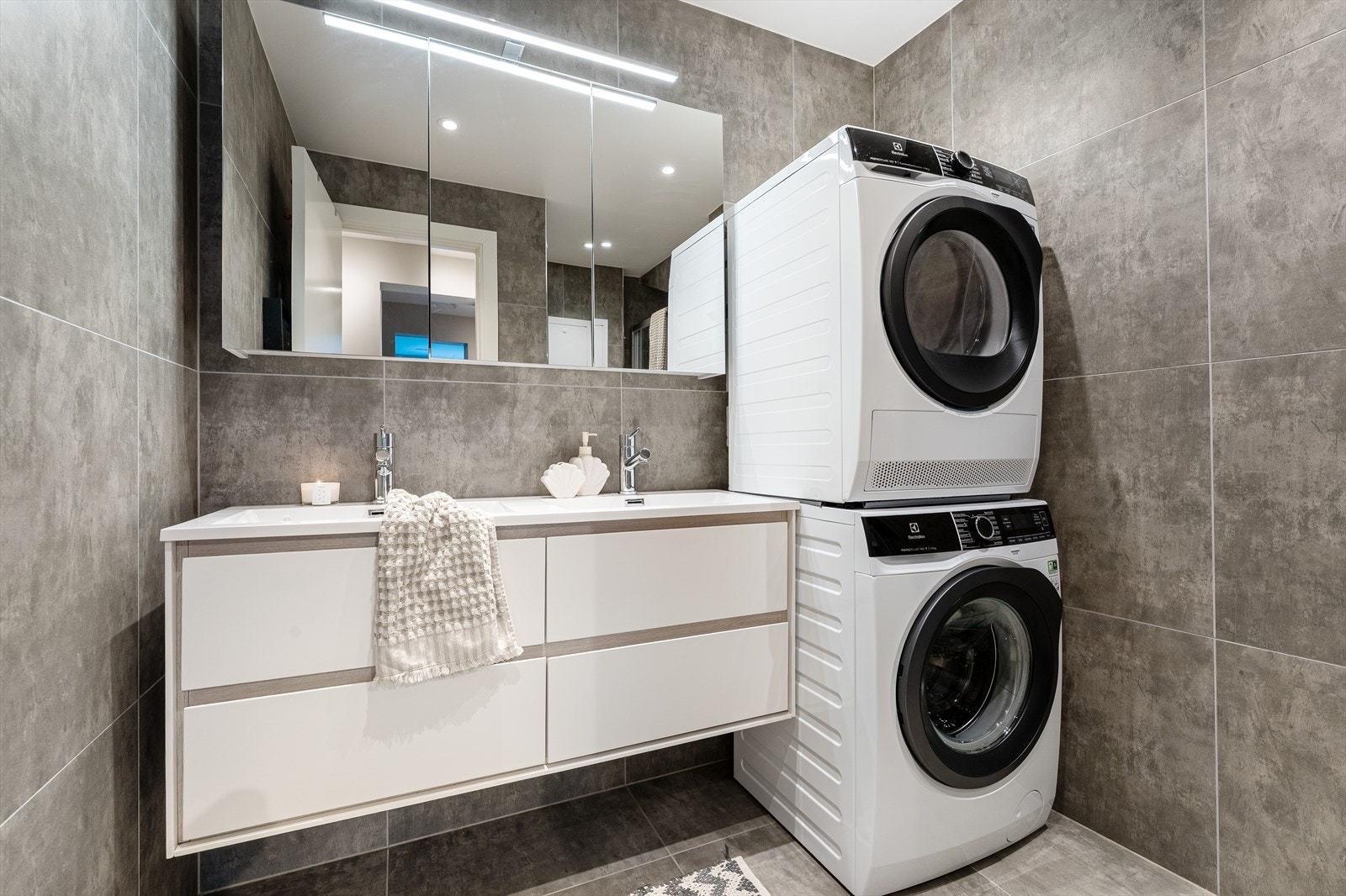 Det er utstyrt med dusjhjørne, mekanisk ventilasjon, vegghengt toalett, baderomsinnredning med servant og belyst speil, samt opplegg for vaskemaskin og tørketrommel Galleribilde