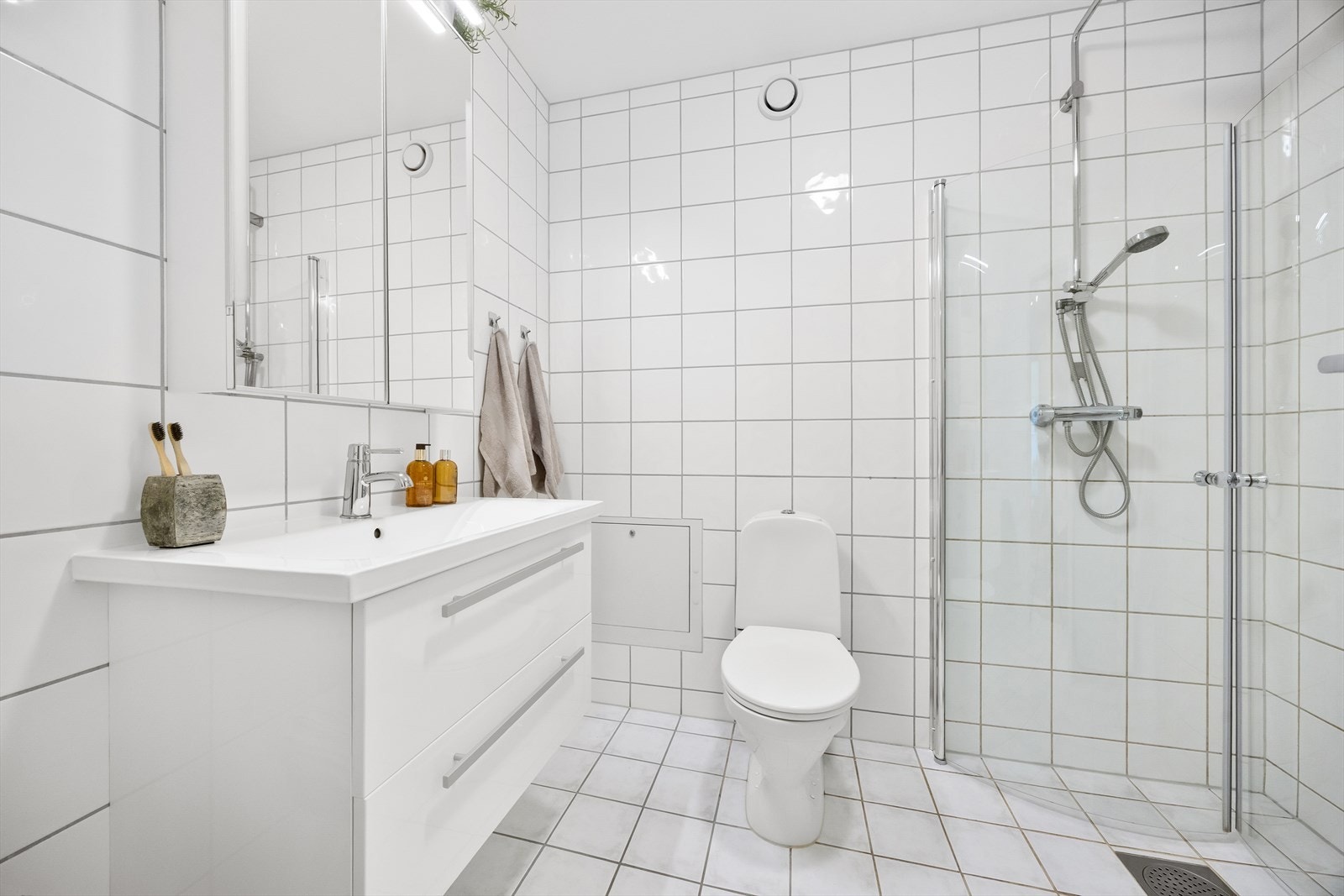 Badet er utstyrt med dusjnisje med innfellbare glassdører, gulvstående toalett og heldekkende servant med skuffer under. Galleribilde