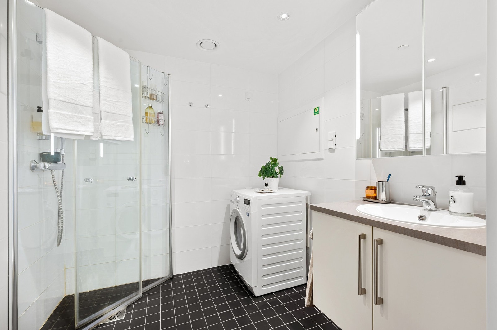 Badet er b.la. innredet med dusjhjørne, veggmontert WC, kran og avløp for vaskemaskin og baderomsinnredning med servant med underskap og speil. Galleribilde