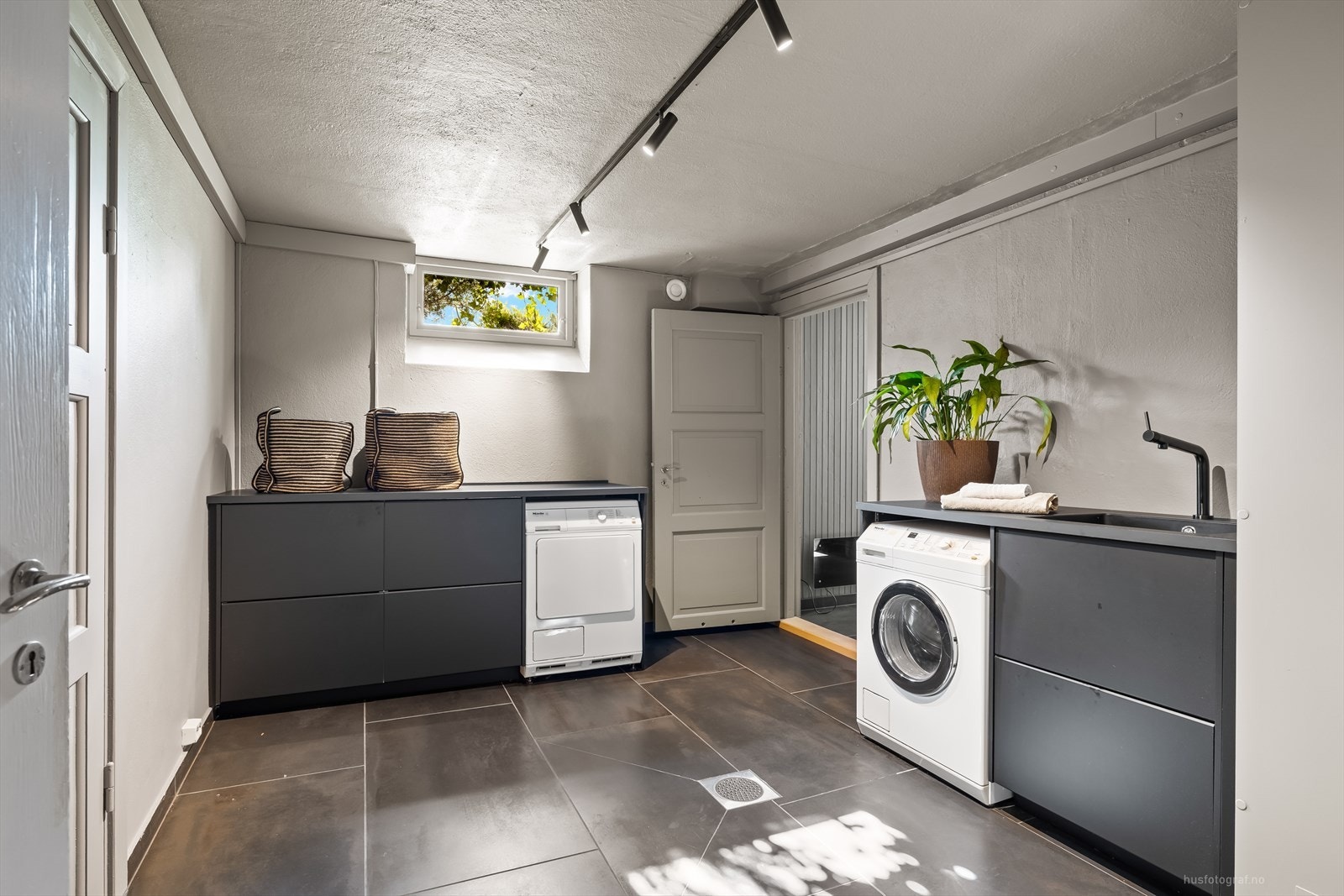 Vaskerommet har flislagt gulv med varmekabler, mørk innredning med skyllekum, benkeplass samt opplegg for vaskemaskin og tørketrommel. Galleribilde