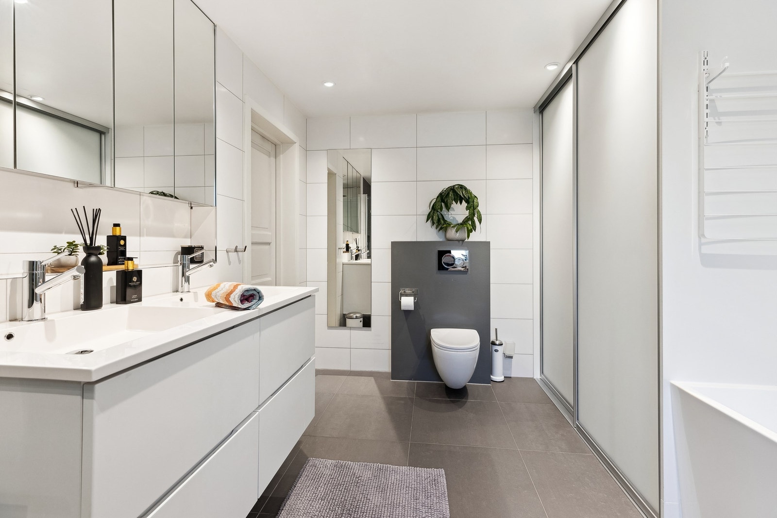 Benkeskap med lyse, glatte fronter med to servanter i helstøpt plate. Romslig dusj 100x90 cm med innfellbare INR dusjvegger i herdet glass, badekar og veggfestet toalett med Geberit sisterne og betjeningsknapper. Galleribilde