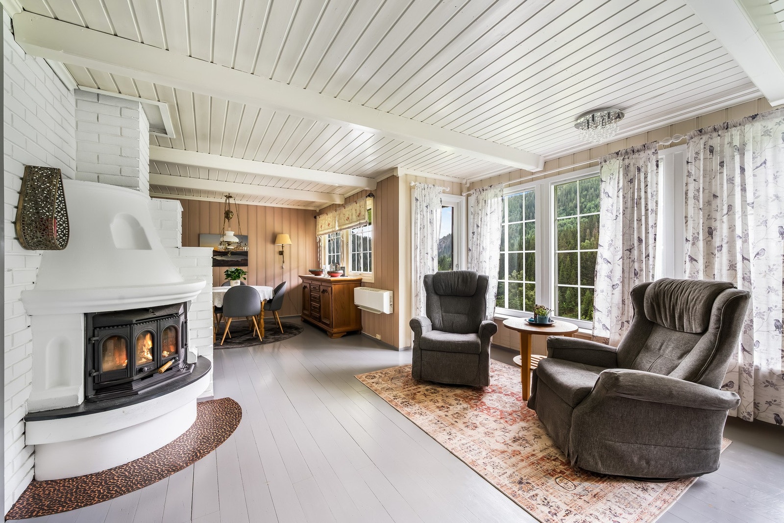Romslig og lys stue med store vindusflater som slipper inn rikelig med naturlig lys og fremhever den flotte utsikten. Planløsningen gir god plass til både spisestue og tv-krok, mens peisen fungerer som et naturlig midtpunkt og bidrar til en lun og innbyde Galleribilde