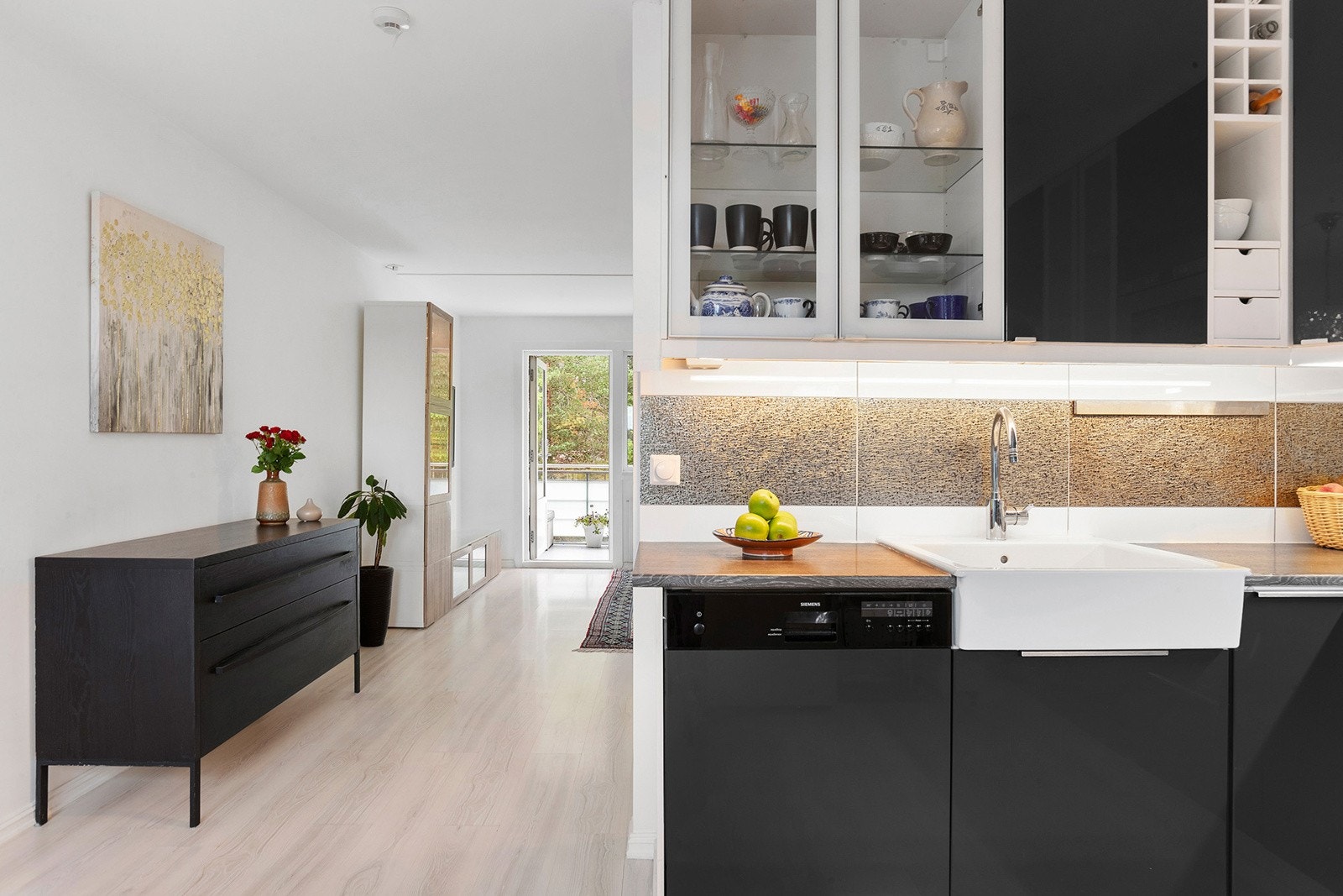 Delvis åpen kjøkkenløsning mot stuen innredet med høyglansfronter, fliser over benk, laminatplate og integrerte hvitevarer. Galleribilde