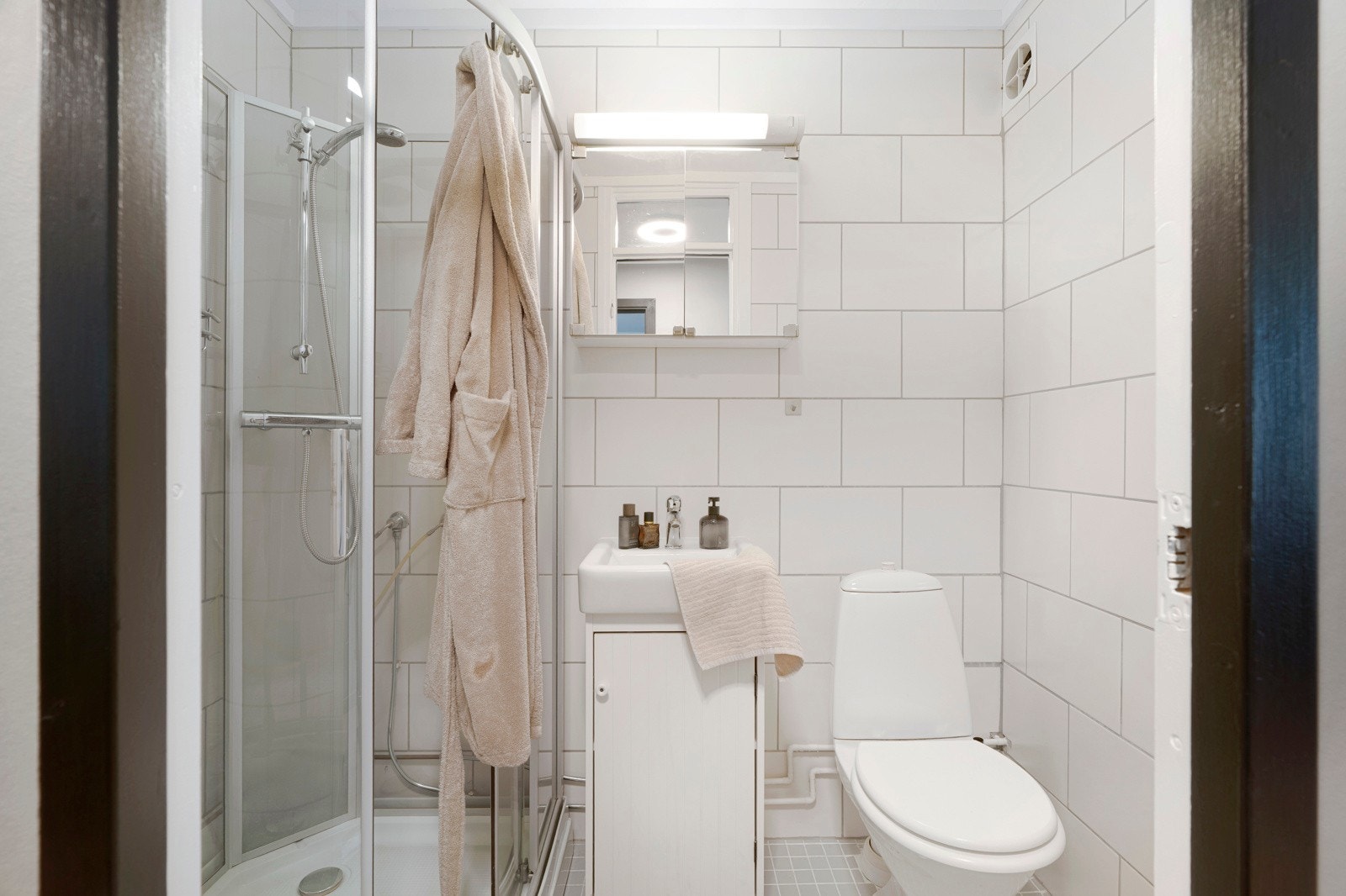Herfra er det adkomst til leilighetens flislagte bad. Badet er lyst og tidløst. Innredningen består av toalett, dusjkabinett, speilskap med belysning og et hvit baderomsmøbel. Galleribilde