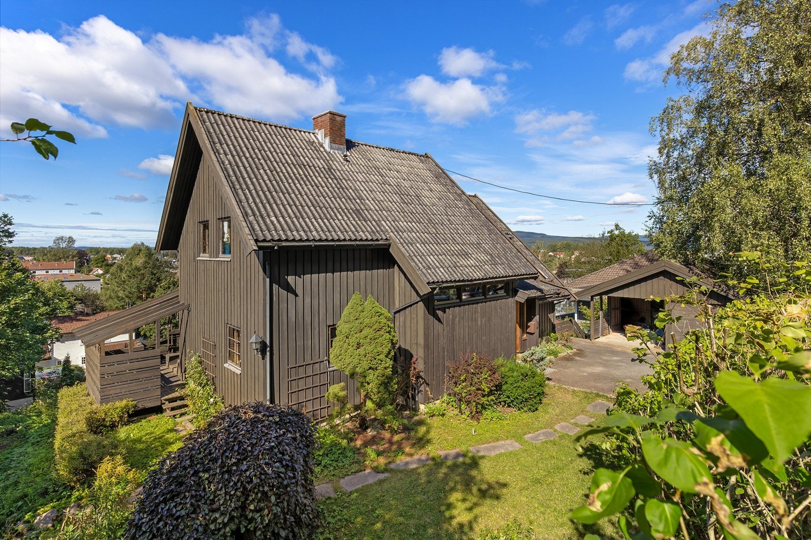 Fasaden sett fra Kurveien. Asfaltert gårdplass, hellelagt gangsti rundt huset til hagesiden. Garasje/carport med lagringsrom under Galleribilde