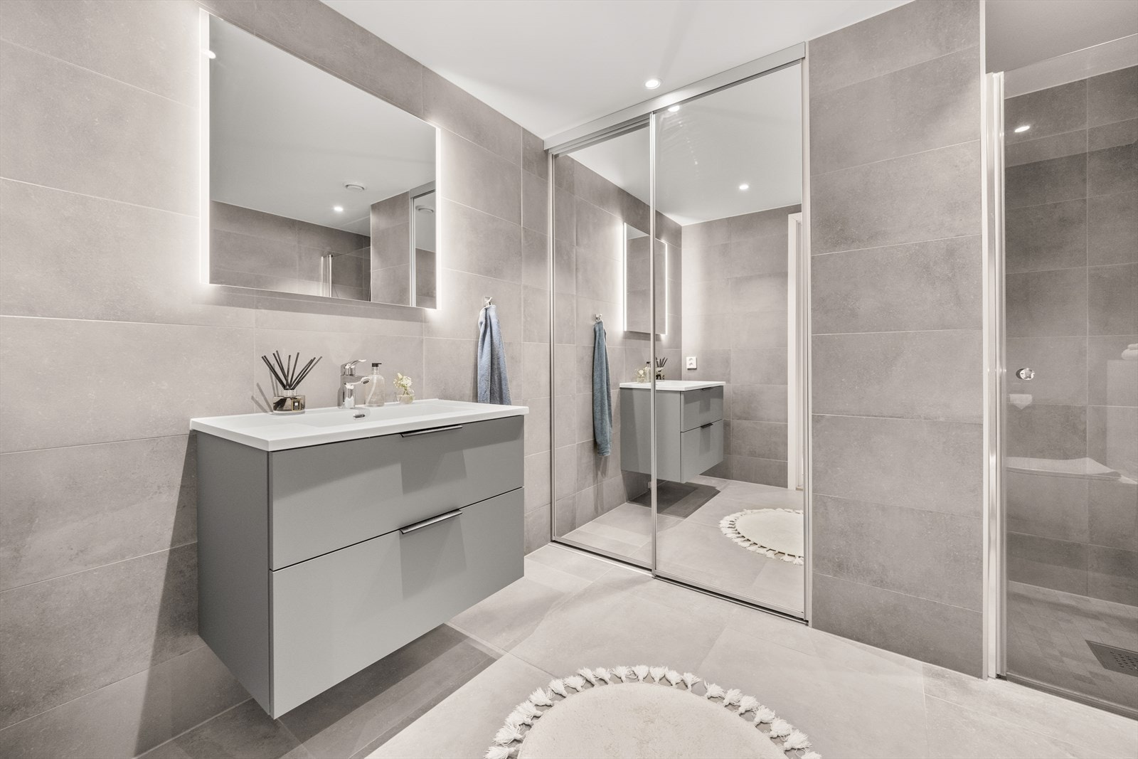 Delikat og pent flislagt bad med opplagg for vaskemaskin og tørketrommel bak skyvedører med speilfronter. Innredningen består ellers av vegghengt klosett, dusjnisje og baderomsinnredning med speil og integrerte ledlys Galleribilde