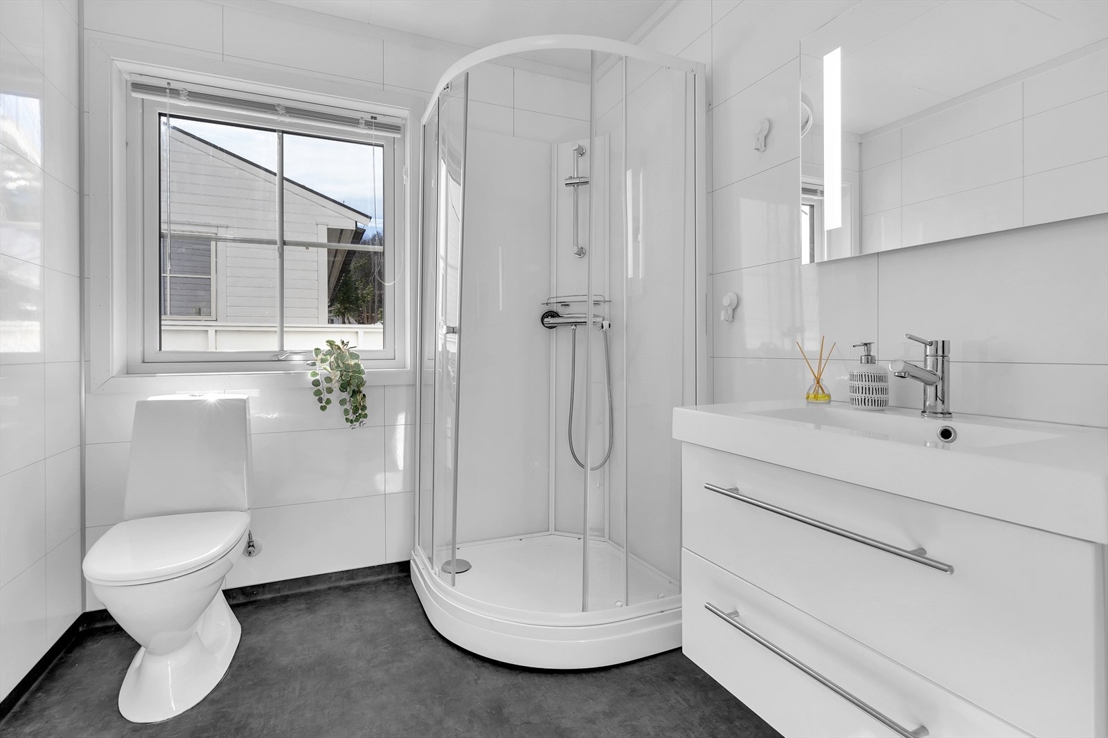 Dette baderommet fremstår som lyst og delikat med praktiske løsninger og moderne detaljer. Det er utstyrt med et dusjkabinett, speil med integrert belysning, servant med underskuffer, et stilrent toalett samt opplegg til både vaskemaskin og tørketrommel. Galleribilde