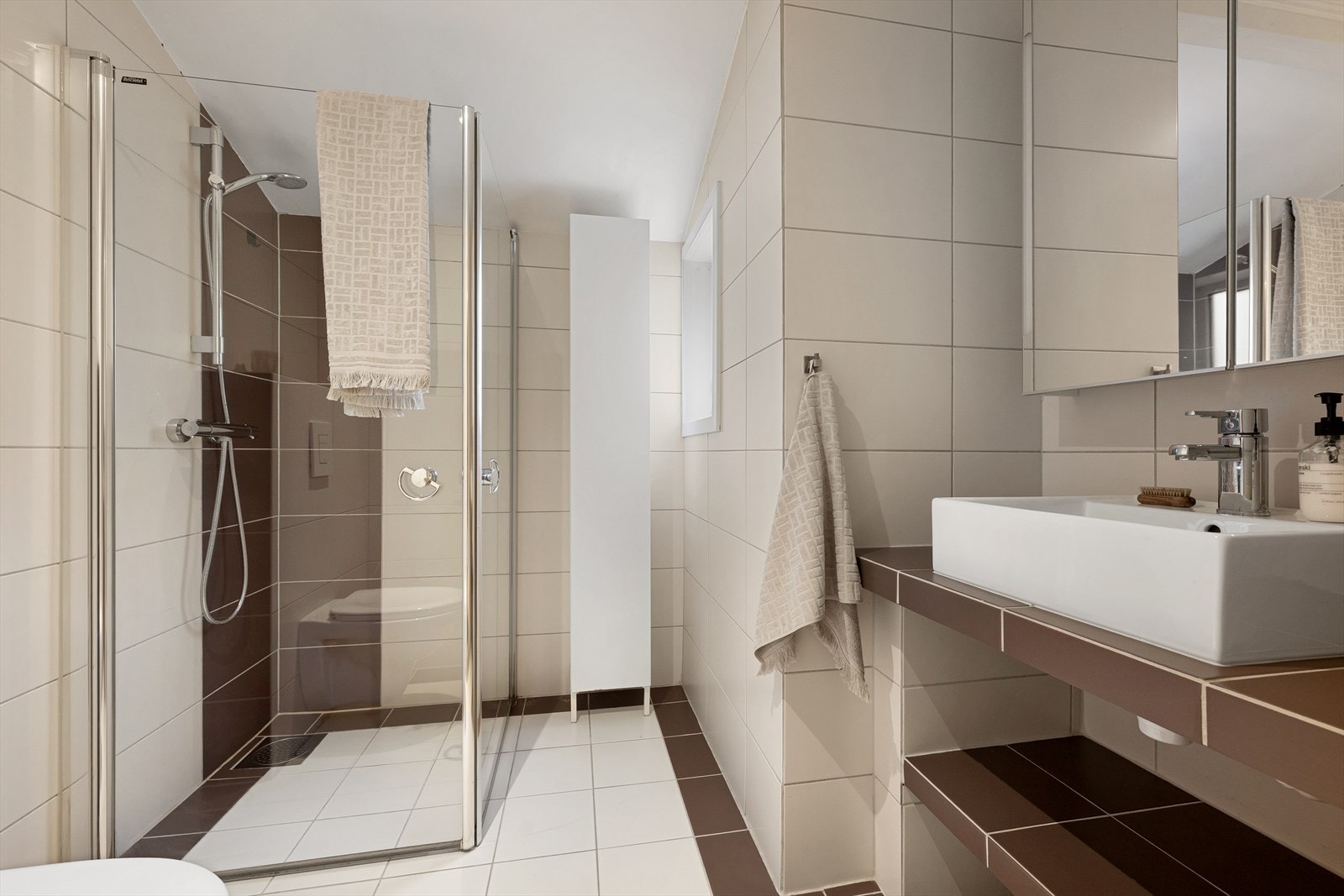 Bad med plassbygget flislagt hylle under servant og veggskap med speilfronter og høyskap med glatte fronter - Dusj med innfellbare dører i herdet glass, servant og veggfestet klosett med innebygget
sisterne Galleribilde