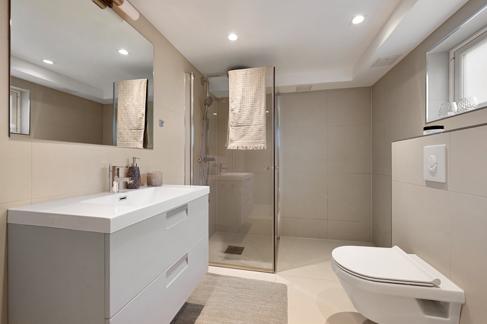 Baderomsinnredning bestående av benkeskap med glatte fronter samt speil og belysning over servant - Dusj med innfellbare dører i herdet glass, servant, veggfestet klosett med innebygget
sisterne og opplegg for vaskemaskin Galleribilde