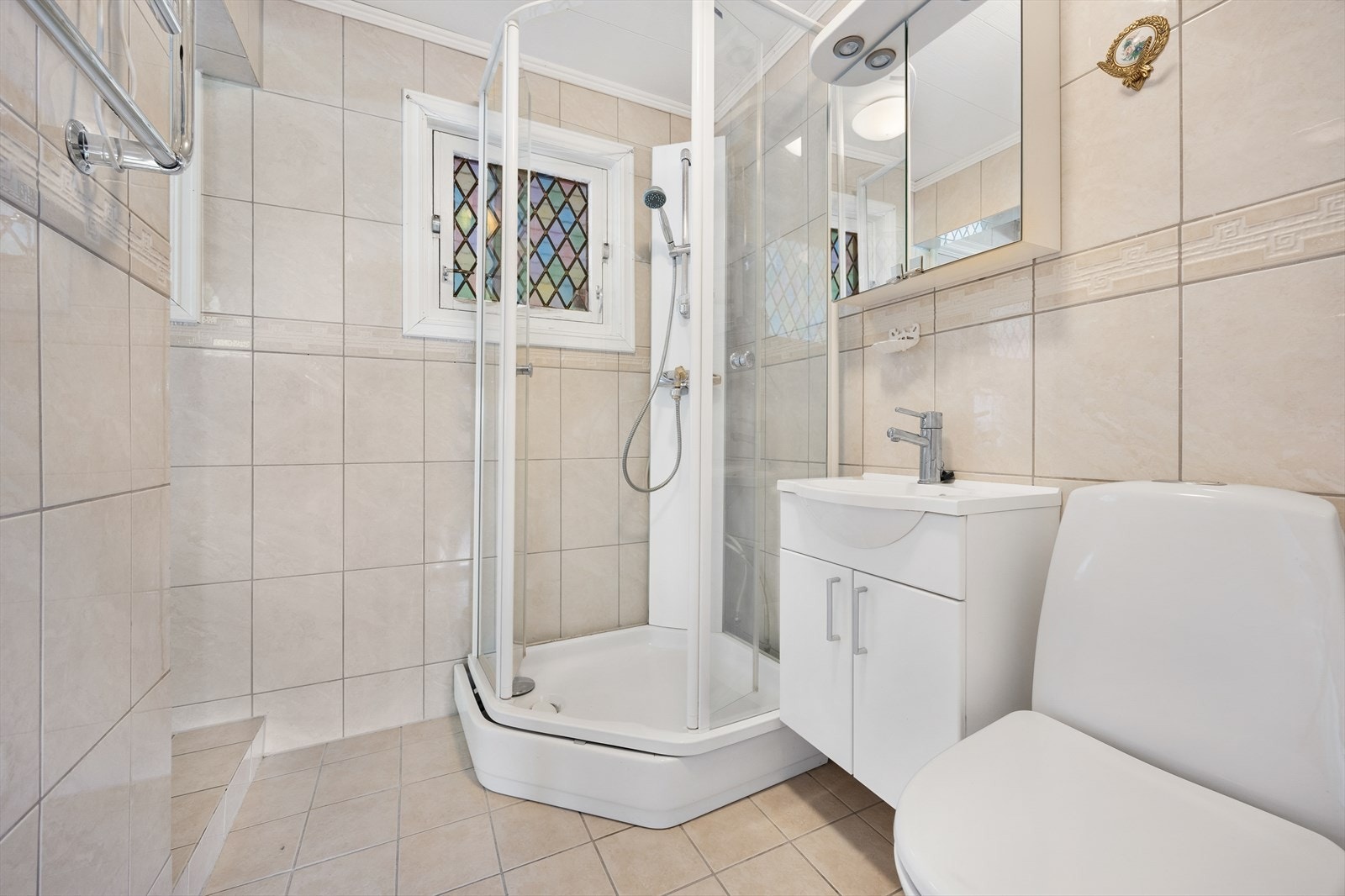 Lyst flislagt bad utstyrt med dusjkabinett, gulvstående wc og servantinnredning med under- og overskap. Galleribilde