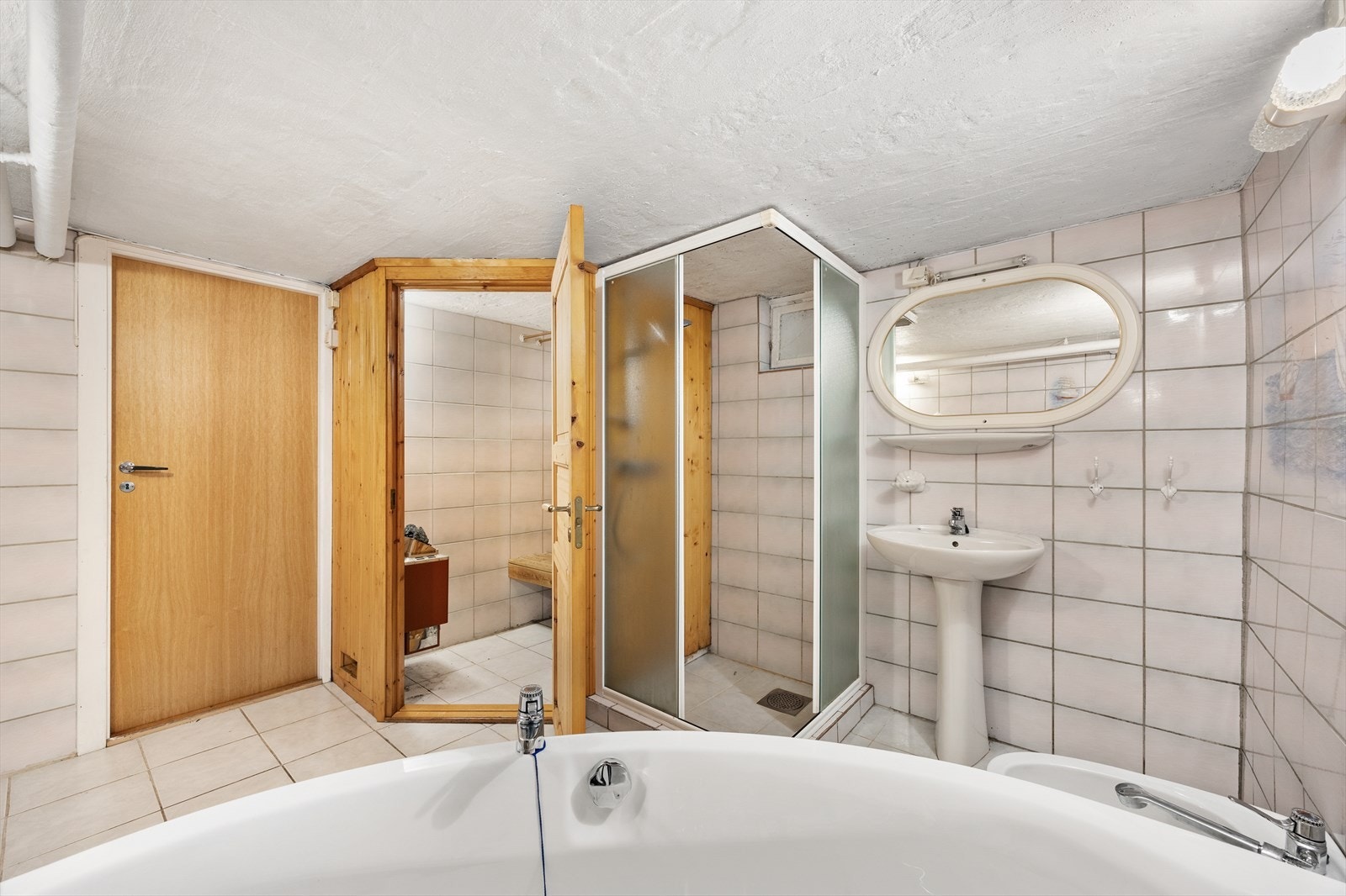 Flislagt bad av eldre standard, utstyrt med hjørnebadekar, dusjhjørne, bide og søyleservant. Galleribilde