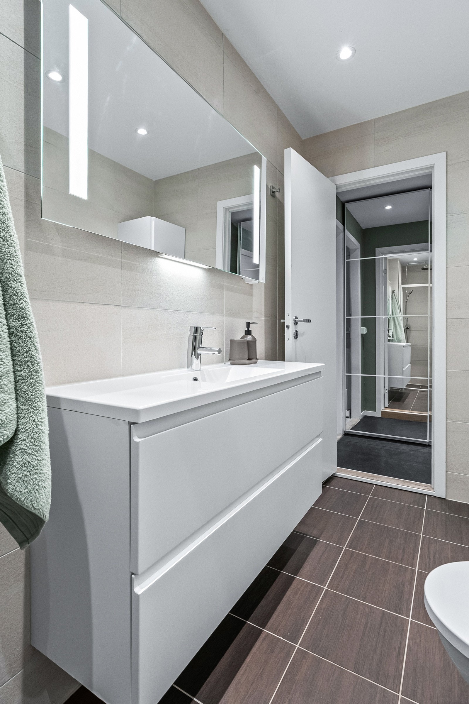 Moderne baderomsinnredning med skuffer og heldekkende servant, stort speil med integrert belysning over. Romslig dusjnisje med glassvegg og dør, både hånd- og regnfallsdusj, samt innebygd hylle for såper. Galleribilde