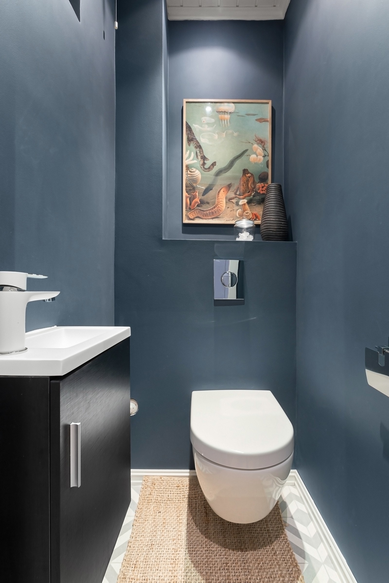 Toalettrommet har samme gulvfliser, glatte, blåmalte veggflater, downlights og gulvvarme. Her er det et veggmontert toalett og en hvit servant på sort servantskap. Galleribilde