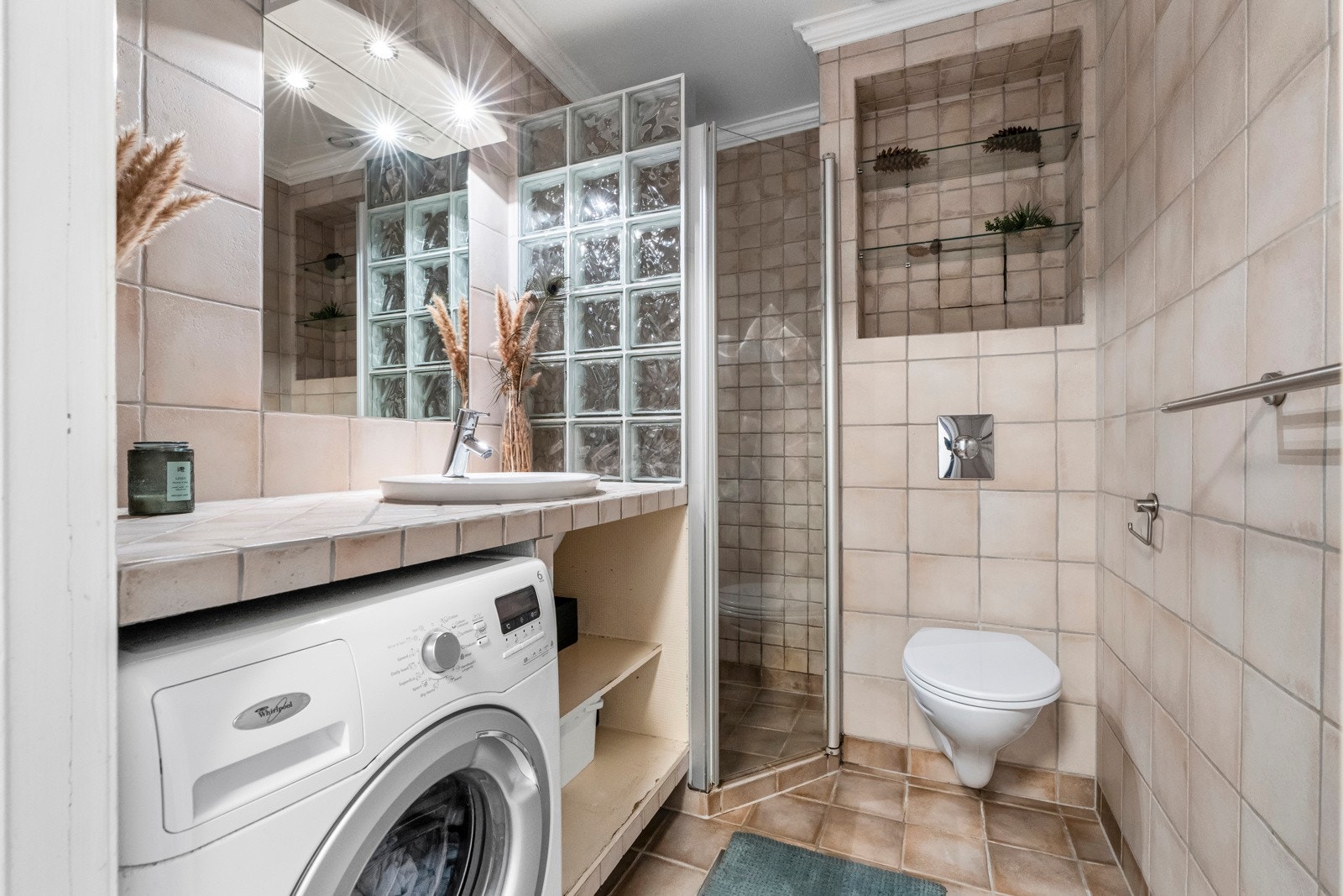 Lyst og flislagt bad som inneholder vegghengt toalett, dusjnisje med dusjdør i herdet glass, plassbygd baderomsinnredning med flislagte flater og nedfelt servant. Galleribilde