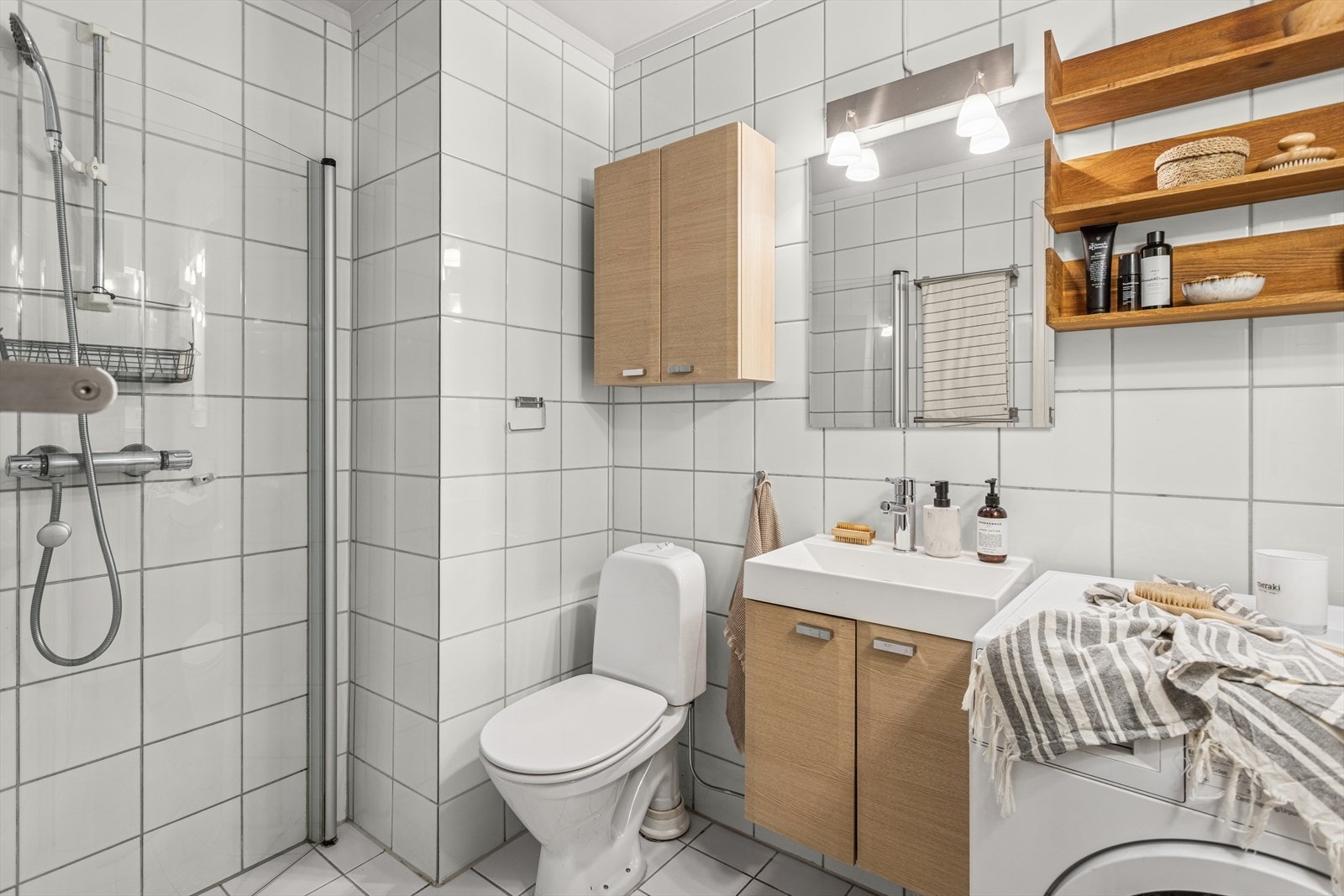 Badet har klassiske hvite fliser på både vegger og gulv. Badet har innredning med nedfelt servant, toalett, dusjvegger/hjørne og opplegg for vaskemaskin. Mekanisk avtrekk med ventil i himling. Galleribilde